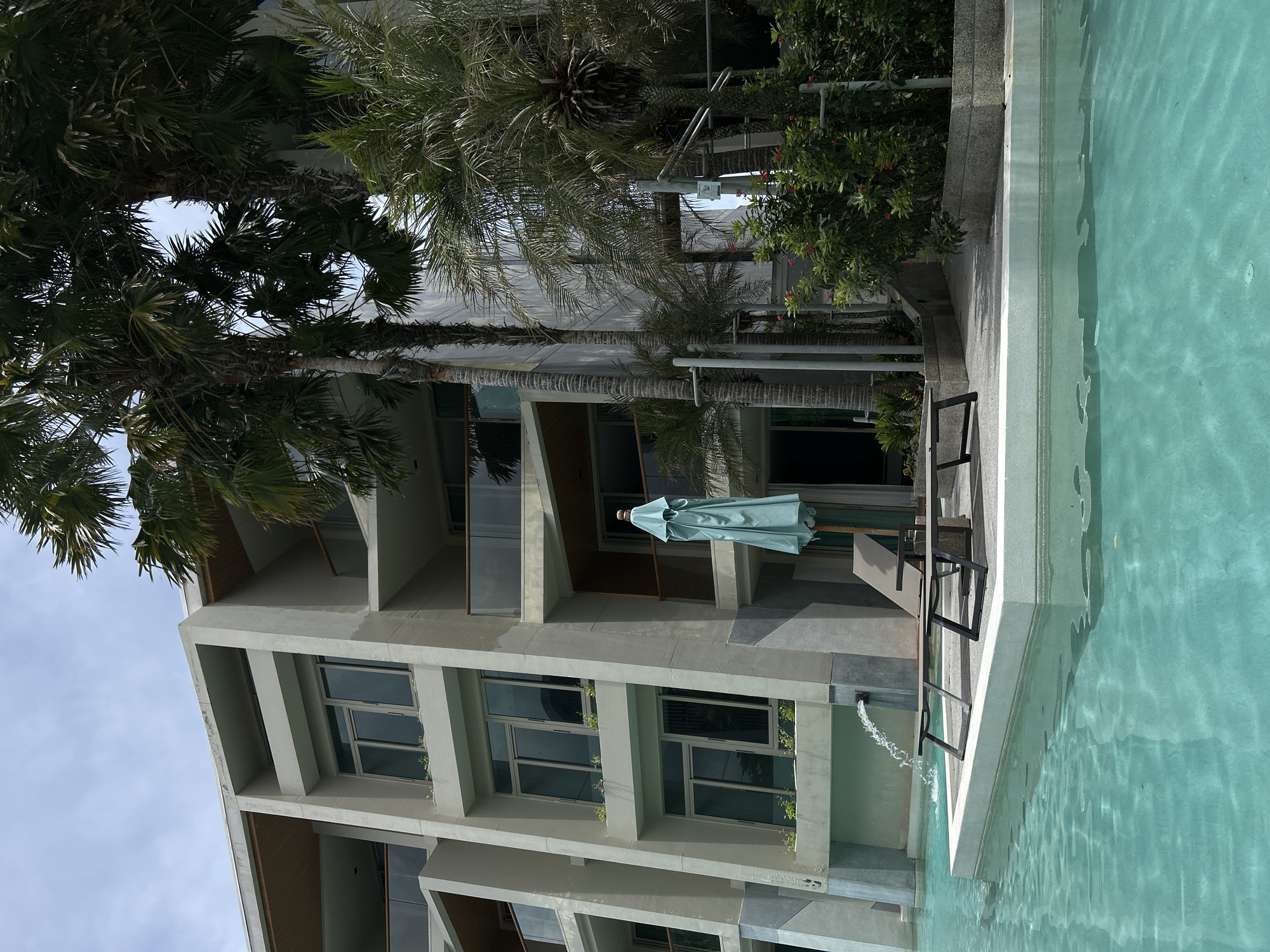 The image shows a multi-story building next to a pool in Rawai, Thailand. The building has a modern architectural style with geometric shapes and large windows. Tropical plants, including palm trees, are visible near the building. A light blue pool umbrella is set up on the terrace between two metal chairs. Water flows into the pool through a fountain along the edge. The sky is partly cloudy.