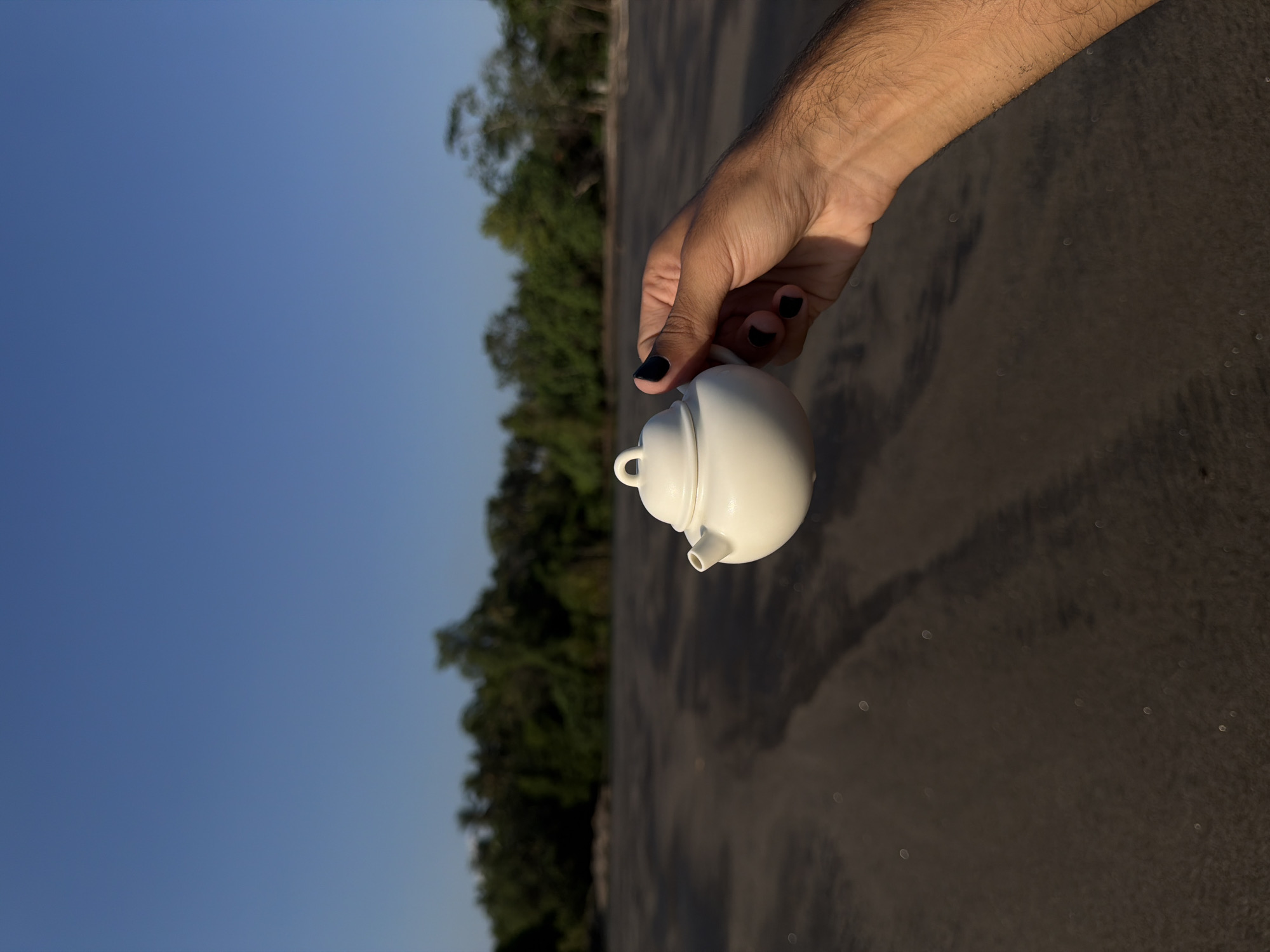 A right hand with medium-brown skin and visible forearm hair, featuring black-painted fingernails, holds a small, white, spherical teapot. The teapot has a short spout and a looped handle on its lid. The subject is positioned in the right midground against a backdrop of dark, possibly wet, sand. Small, out-of-focus circular light reflections are present on the sand surface. In the background, a dense line of out-of-focus green foliage and trees stretches horizontally. The upper portion of the image is dominated by a clear, bright blue sky with no visible clouds. The scene is located in Selemadeg Timur, Indonesia.