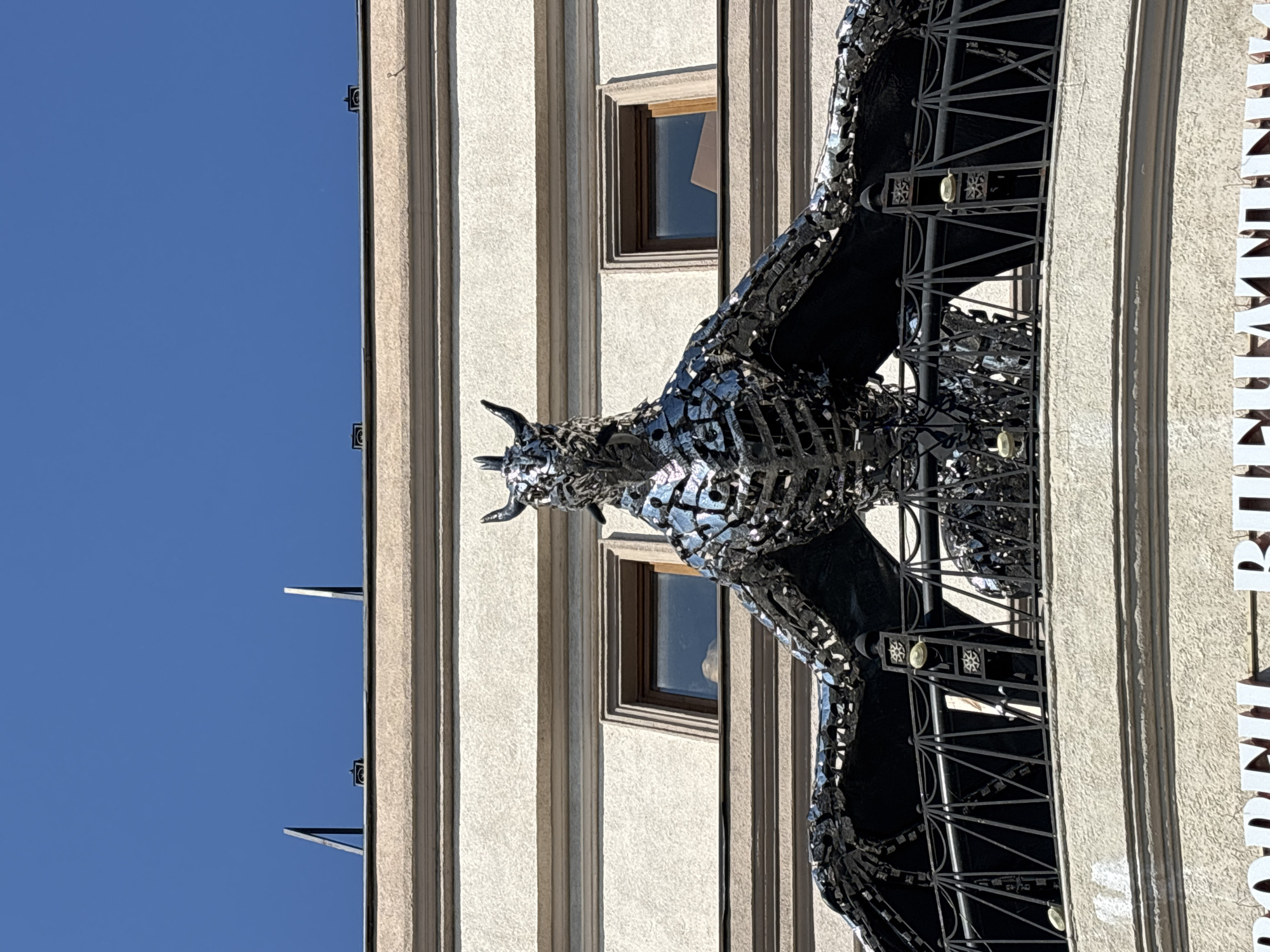 An ornate, metallic sculpture of a dragon with outstretched wings is perched on a balcony atop a building in Kazan, Russia. The dragon, constructed from numerous reflective pieces of metal, has a horned head and a segmented body. The building itself is beige with two visible windows and a textured facade. The balcony railing is black and features decorative metalwork. The sky above is a clear, bright blue. The bottom of the image shows a portion of a white sign with dark lettering, partially obscured.