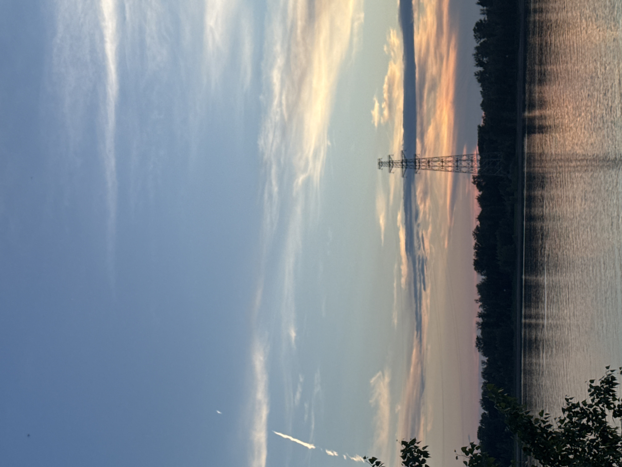 The image shows a wide river in Noviy, Russia, at dusk. The sky is a gradient of blue, with wispy clouds tinged with orange and pink from the setting sun. A solitary, tall electrical tower stands on the far bank, silhouetted against the colorful sky. The dark line of trees forms the horizon behind the tower and stretches across the frame. The surface of the river reflects the sky's colors and the distant trees. In the foreground, the dark leaves of a tree branch are visible on the left side of the image. There are no people or animals visible in the scene.