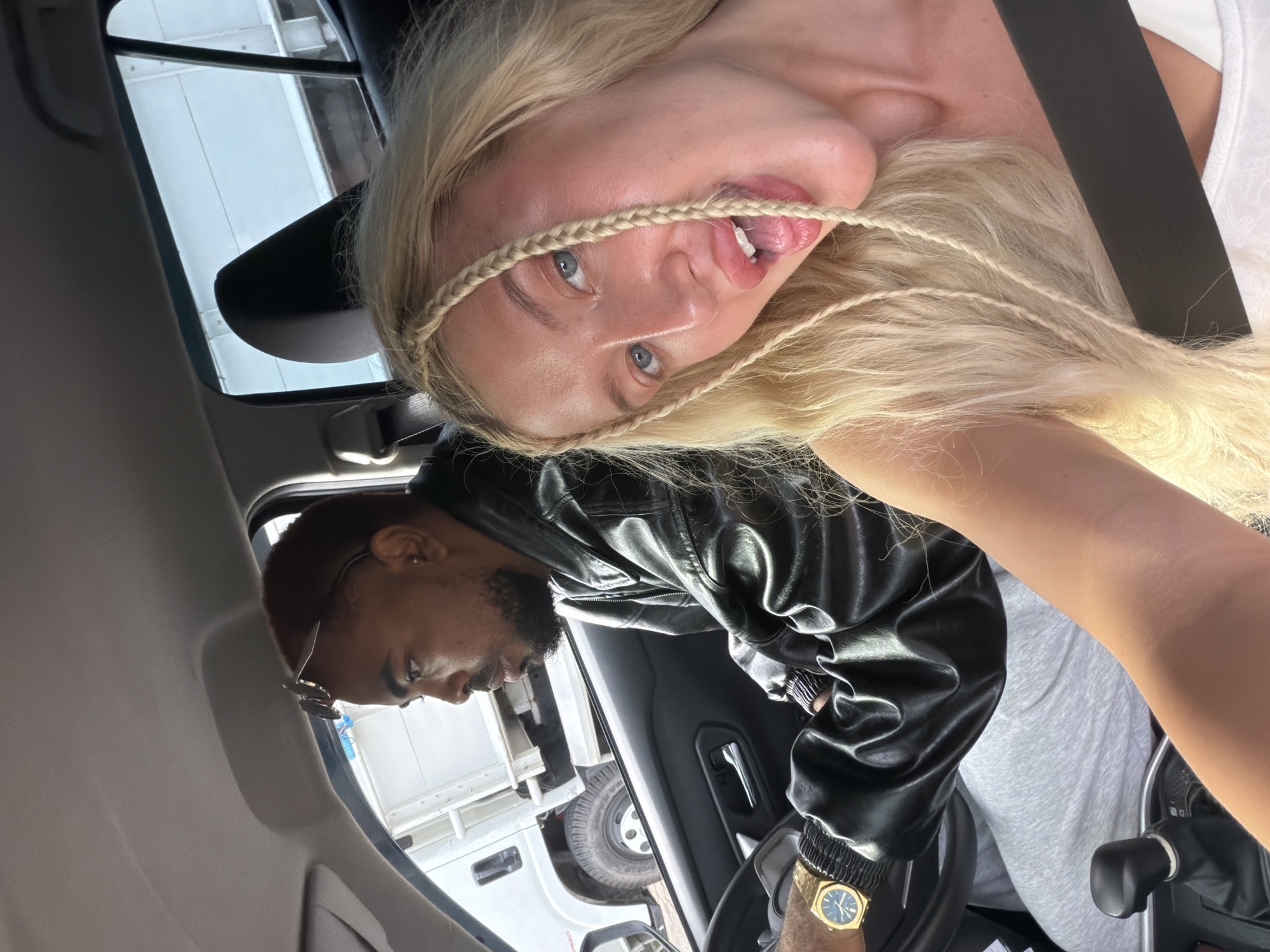 In the interior of a vehicle, a young Caucasian woman with blonde hair styled in braids is in the passenger seat, her face close to the camera, with her mouth open and her tongue slightly extended. She is wearing a white top and a black seatbelt. The driver, a Black male with short, reddish-brown hair and a beard, is looking to his left, out the window. He is wearing a black leather jacket and a gold watch on his left wrist. The vehicle's interior includes a black dashboard and a grey gear shifter. Outside the car, a white truck is visible through the window. The scene appears to be in the city of Mengwi, Indonesia, based on the provided location information.
