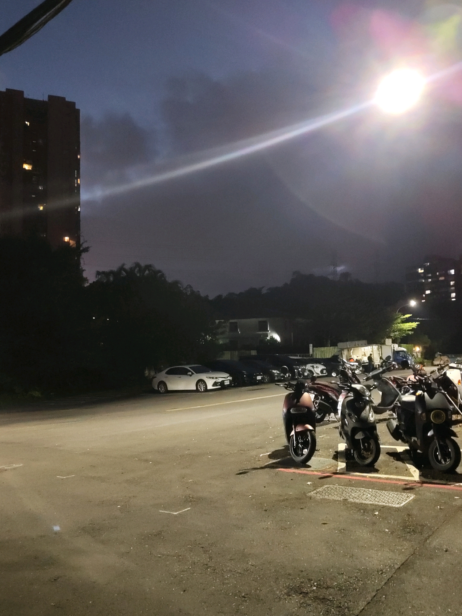 The image captures a street scene in New Taipei City, Taiwan, during twilight or night. In the foreground, a paved road is visible with several parked scooters, including a maroon one on the left, a black one in the middle, and a darker one on the right. Some of these scooters are parked near metal manhole covers and a red and white striped border. Further down the street, a white sedan is parked. To the left of the road, a tall, dark-colored residential building with illuminated windows stands prominently. Behind the building and the road, a dense area of trees and foliage is present. In the background, a dimly lit hillside is visible, shrouded in what appears to be mist or clouds, with a bright, diffused light source illuminating the sky and casting a distinct beam. Another multi-story building is visible on the far right of the image, also with lit windows. The overall atmosphere is dark and somewhat hazy, with artificial lights from buildings and the sky contributing to the illumination.
