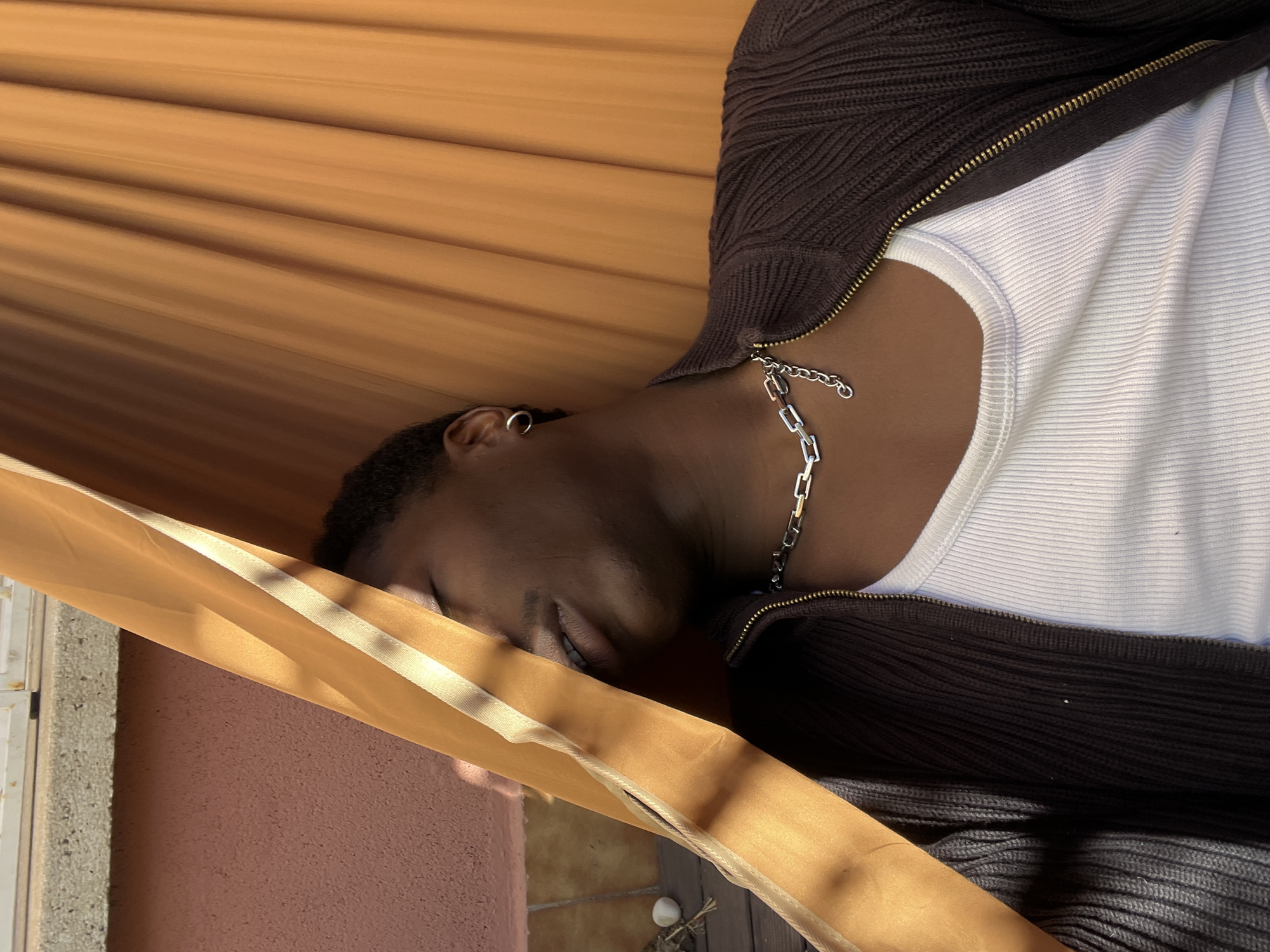 A person with short, dark hair, a silver hoop earring in their left ear, and a silver chain necklace is visible in the image. They are wearing a white ribbed tank top and a dark brown ribbed cardigan with a zipper. The scene is brightly lit, likely by natural sunlight.
The person is partially obscured by a golden-colored drape with a light-colored ribbon edge, suggesting a window or doorway setting. Behind the drape is a peach-colored wall and a white windowsill. Below, a portion of tiled flooring is visible, with a decorative object (possibly a small, round stone) on the floor. The image is vertically oriented, implying it was taken on a mobile phone. The overall impression is that the photo was taken indoors, near a window with a decorative drape.
There is no clear evidence in the image that indicates the location is in Barcelona, Spain.