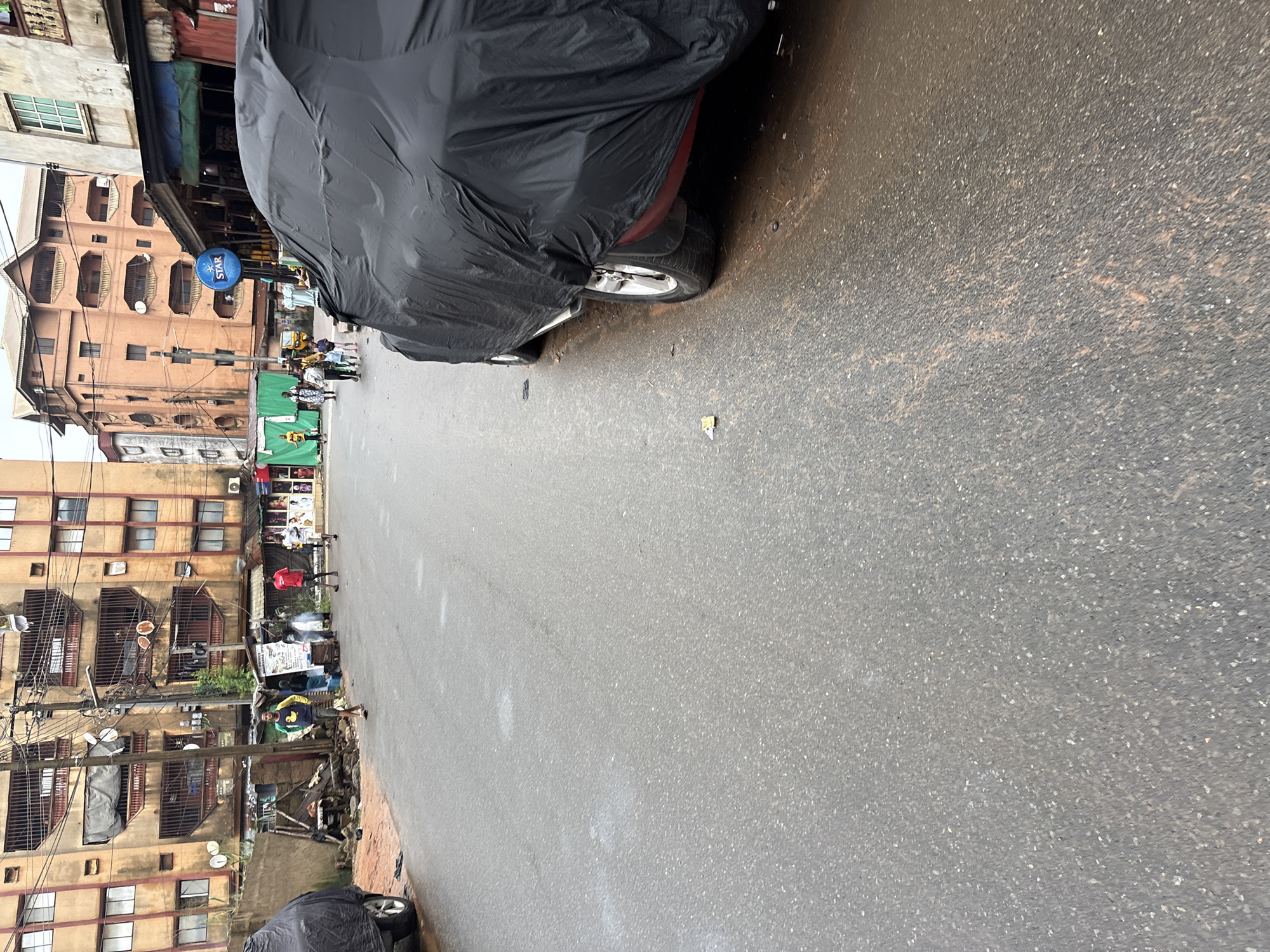 The image depicts a street scene in Onitsha, Nigeria, likely during or after a rain shower, as evidenced by the wet asphalt and reflective surfaces. In the foreground, a car is covered with a black tarp, with its rear wheels visible. The street is a wet, grey asphalt road with some scattered debris.
Along the side of the road, there are several buildings, the most prominent being a multi-story structure with a weathered, yellowish-orange facade, featuring numerous balconies with metal railings. To the left of this building, a smaller, rougher structure with corrugated metal roofing and exposed wooden beams is visible. Further down the street, another multi-story building with a salmon-pink hue and a distinctive pitched roof is present, along with other urban structures that appear to be shops or residential buildings.
Several people are present in the scene. On the left side of the street, a young man in a green and yellow shirt and dark shorts is walking. Further down, a group of people, including individuals in red and white shirts, are interacting or walking. In the middle distance, a cluster of people are gathered near a stall or shop. In the background, a few more individuals are seen walking or standing. A small, three-wheeled vehicle (likely a tuk-tuk or auto-rickshaw) is parked on the side of the road. A blue circular sign with a white star and the word "STAR" is prominently displayed above one of the shops. The overall impression is of a busy, everyday urban environment in Onitsha.
