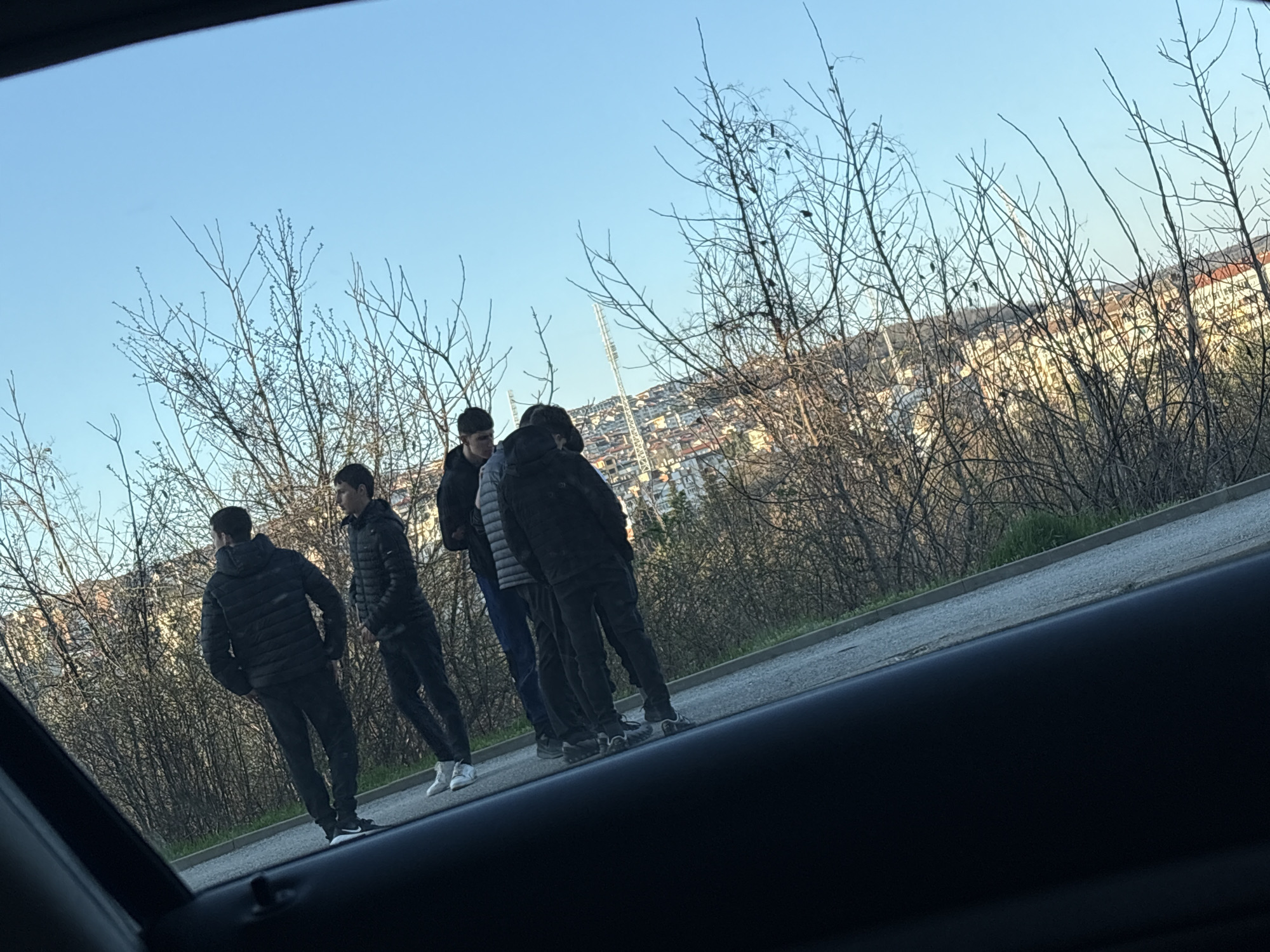The image shows five young men standing on a roadside in Veliko Tarnovo, Bulgaria. The men are all dressed in casual clothes. A background shows a cityscape with buildings and what appears to be a stadium light tower. The photo is taken from inside a car, looking out through the window. The weather appears to be clear and sunny.