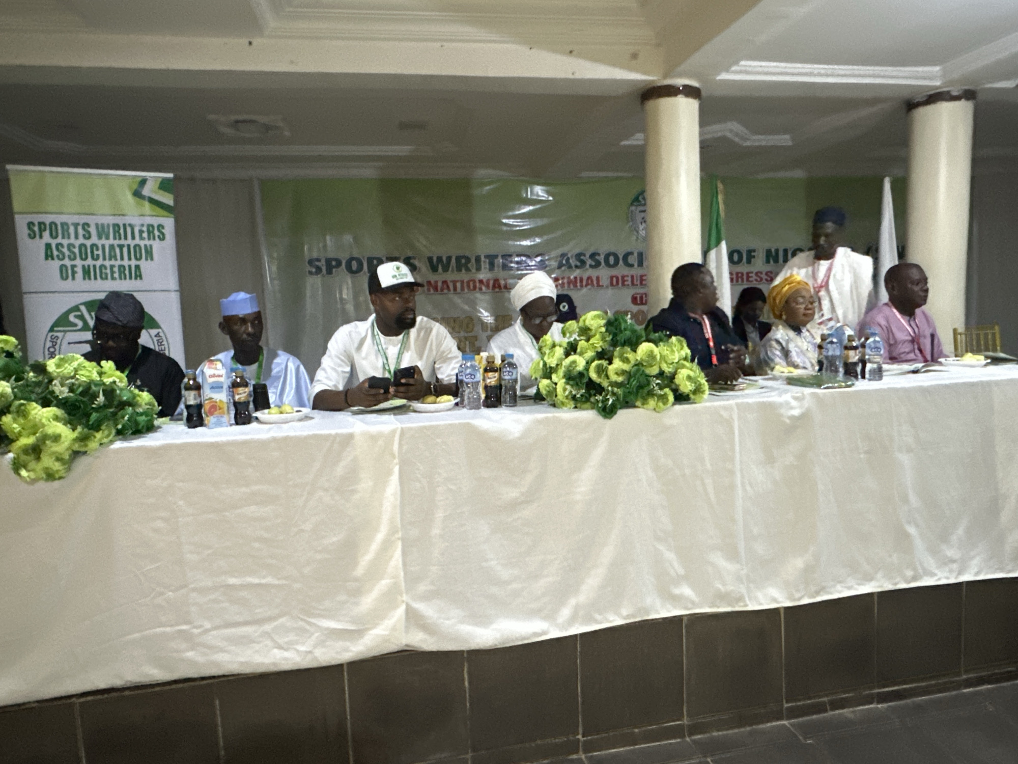 The image depicts a formal event, likely a conference or a meeting, taking place in Barden Yana, Nigeria. Several individuals are seated at a long table covered with a white cloth, facing the camera. Behind them, a banner displays "SPORTS WRITERS ASSOCIATION OF NIGERIA".
The individuals seated at the table are dressed in various attires, including traditional Nigerian clothing and formal shirts. Some are wearing caps, and one person is wearing a yellow headscarf. There are bottles of beverages and some food items on the table.
In the background, additional banners and flags are visible. The overall environment appears to be an indoor venue with overhead lighting. The people are engaged in what seems to be a seated presentation or discussion, with some individuals looking towards the camera. The scene suggests an organized and official gathering related to sports journalism in Nigeria.