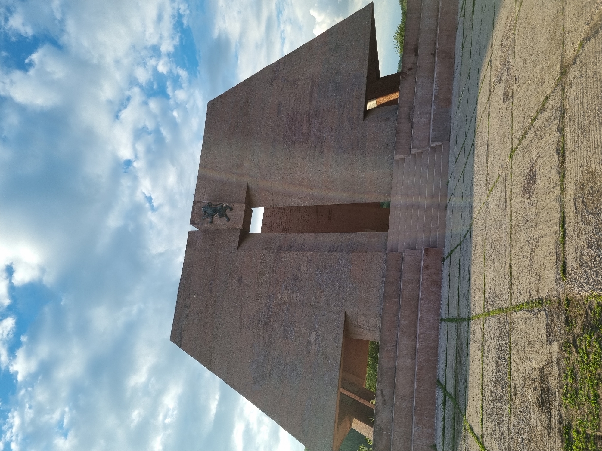 The image shows a monumental structure in Gurgulyat, Bulgaria. The structure is made of a reddish-brown concrete or stone material and features a large central block with a cross-shaped opening. A bronze lion emblem is mounted on the left side of the block. A series of steps lead up the right side of the structure. The ground is paved with large concrete slabs with grass growing in the joints. The sky is visible in the background, filled with fluffy white clouds. The photo is taken at a low angle, looking upwards at the structure.
