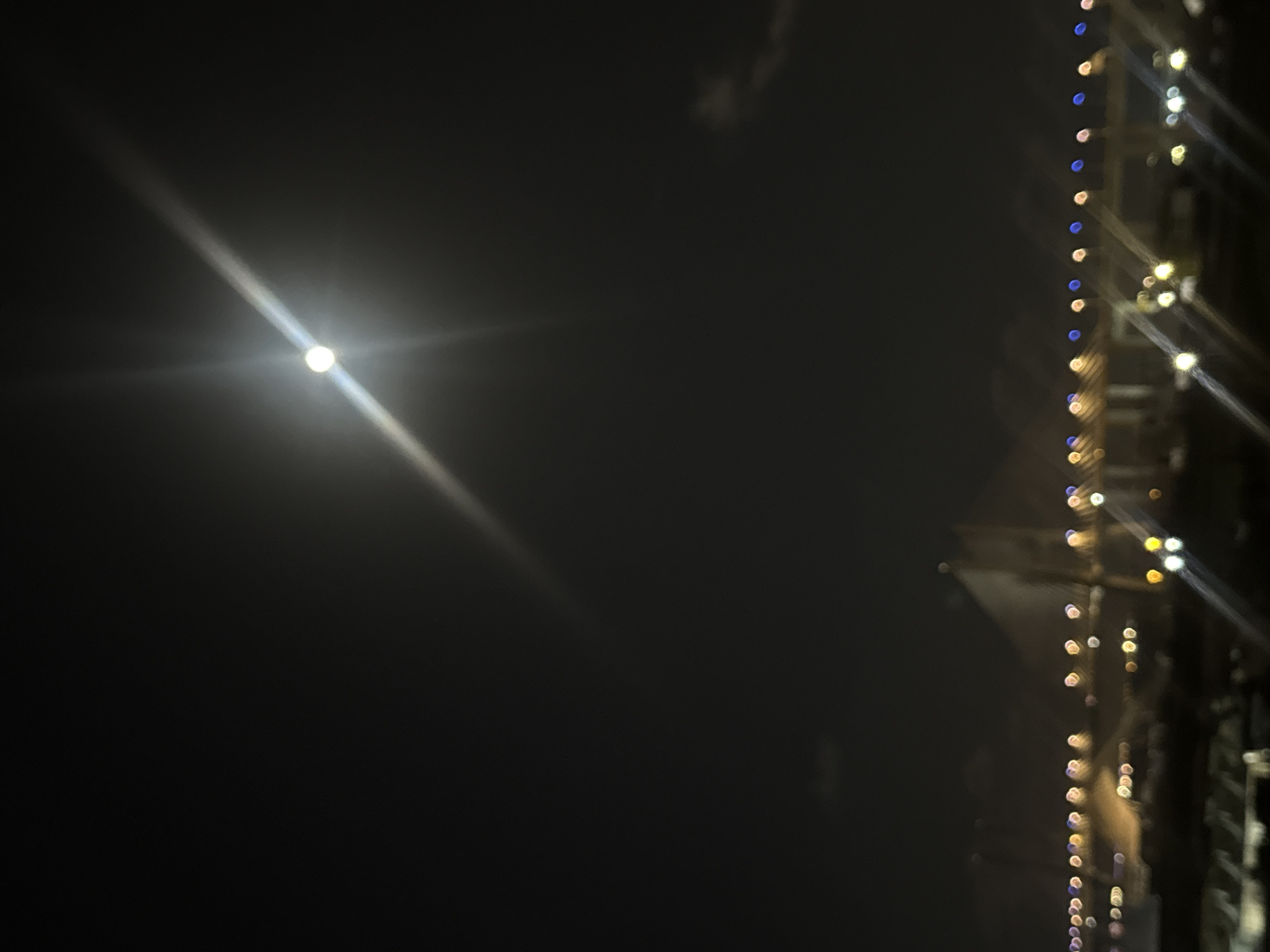 This nighttime image, captured in Loures, Portugal, presents a dark sky illuminated by distinct light sources. On the left side, a brilliant, circular light source, possibly the moon or a powerful lamp, emits a prominent diagonal lens flare, stretching across the frame. Hints of faint, wispy clouds are discernible in the upper-middle section of the dark sky.
To the right, a vertical series of blurred, circular lights, likely decorative string lights, adds a touch of color to the scene. These lights display hues of blue, yellow, and white, creating a bokeh effect against an indistinct, dark background that suggests a building or structure. The overall impression is one of a quiet night, with the bright celestial or artificial light contrasting with the festive glow of the decorative lights. No people or visible text are present in the image.