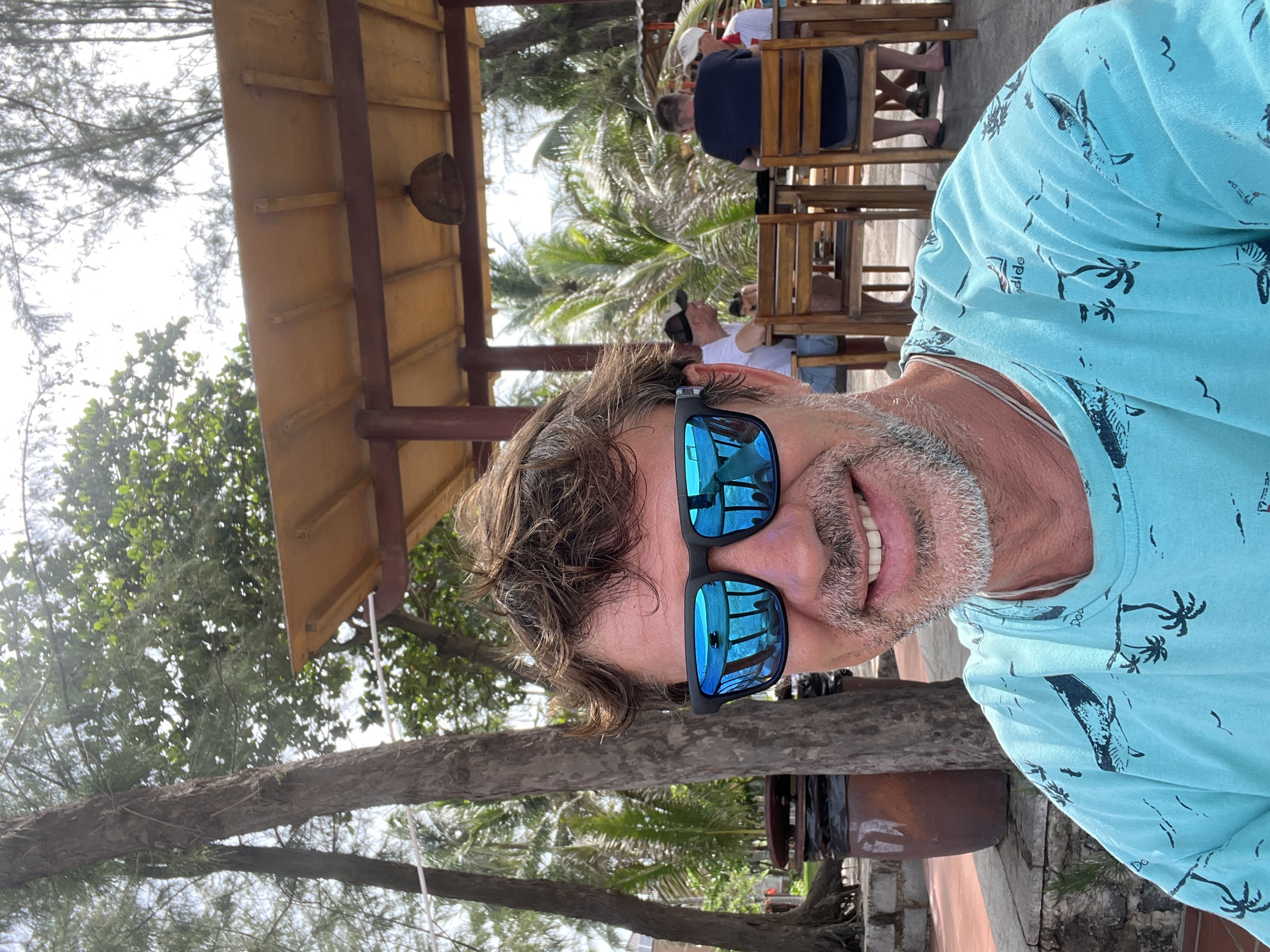 A medium shot shows a man in sunglasses and a teal t-shirt with a sea creature print. He is smiling and looking directly at the camera. He has short, messy brown hair and a grey beard. The man is outdoors, with trees and a thatched-roof structure visible in the background. The setting appears to be a beachside cafe in Mũi Né, Vietnam, as indicated by the palm trees and tropical foliage. Several other people are seated at tables in the background, suggesting a public space. The lighting is bright, indicating daytime.