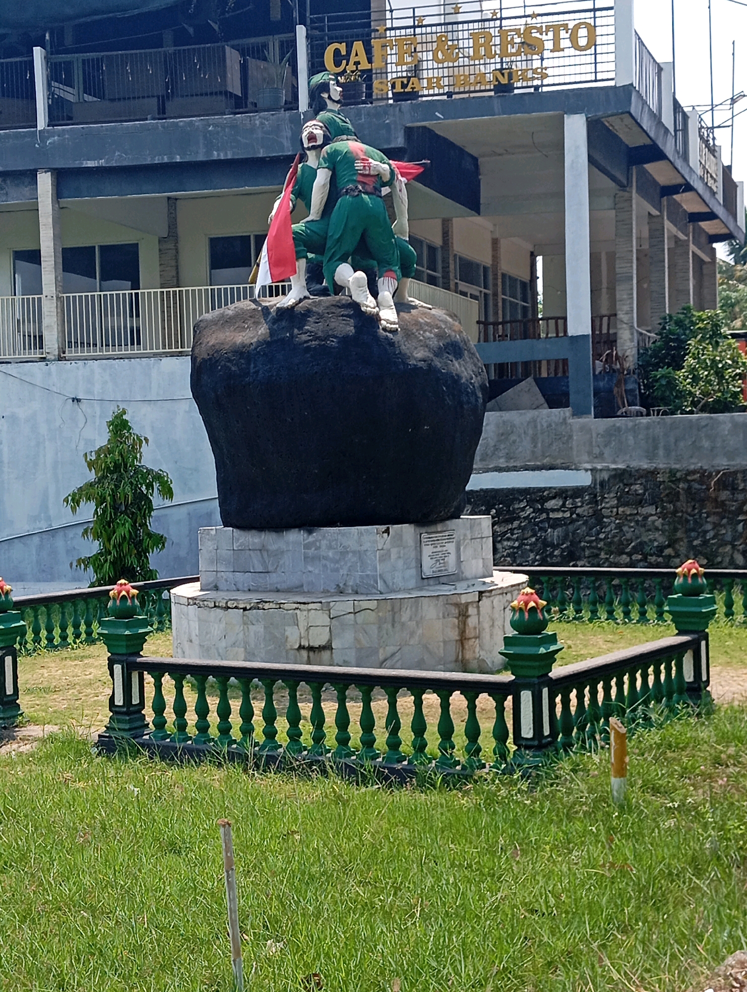 The image shows a statue of two figures, likely soldiers, in a defensive posture on a large rock. One figure is carrying a flag, possibly the Indonesian flag, which is red and white. Both figures are wearing green uniforms. The statue is situated in front of a multi-story building with the words "CAFE & RESTO" visible on the upper facade. The building appears to be an establishment with balconies and dining areas. The statue is surrounded by a low, ornate green fence with decorative elements at intervals. The ground in front of the fence is covered in green grass, and there is a small, slender tree to the left of the statue. The environment suggests an outdoor public space, possibly a park or a plaza adjacent to the cafe. The overall scene depicts a commemorative or historical monument, likely in a location within the city of Leman Benawi, Indonesia, given the context of the prompt.