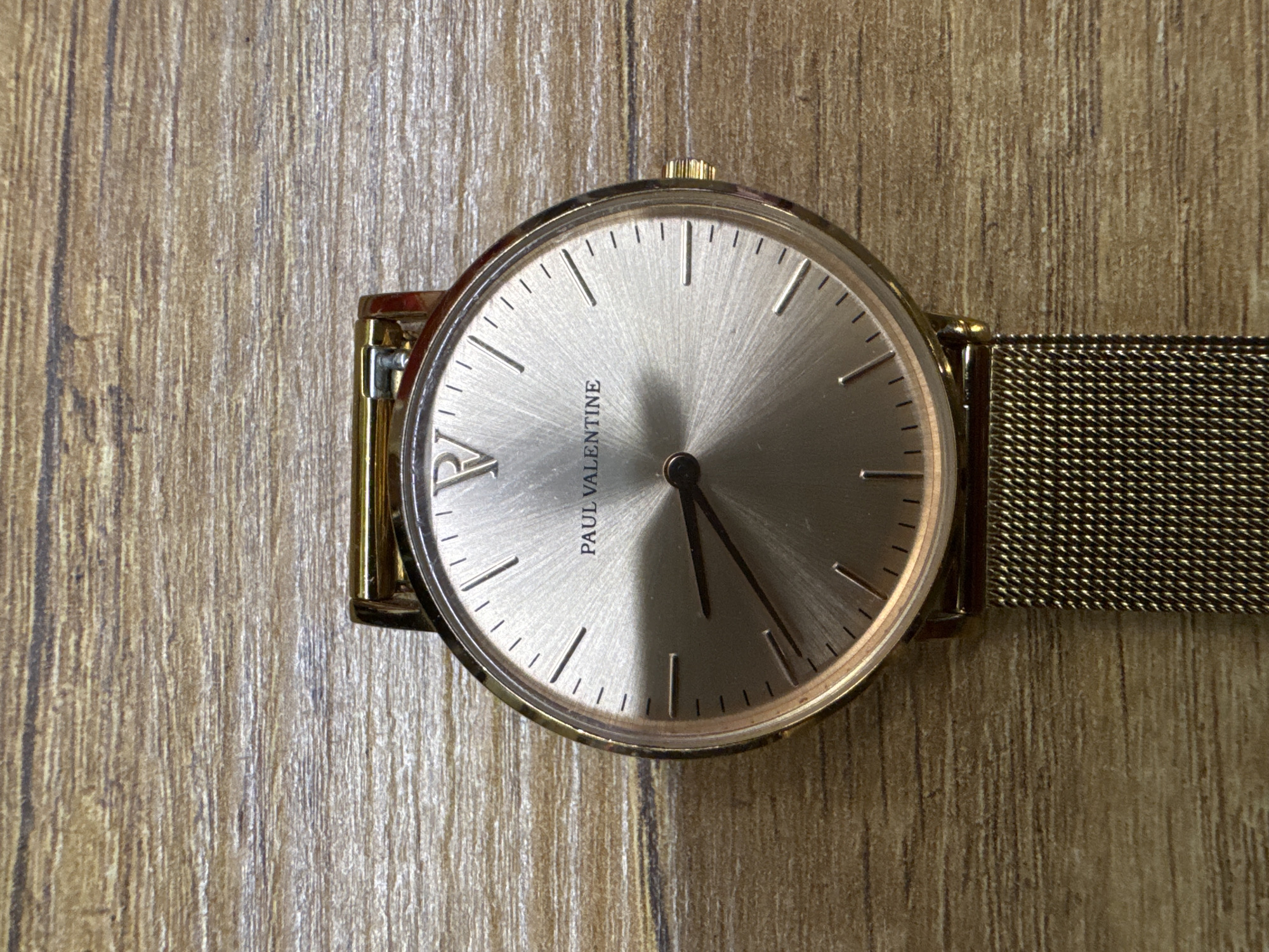 A wristwatch sits on a wood-grain surface. The watch face is silver with a golden frame. The brand name "PAUL VALENTINE" is printed in black on the dial. The watch has a mesh band made of a similar gold material. The minute and hour hands are black.