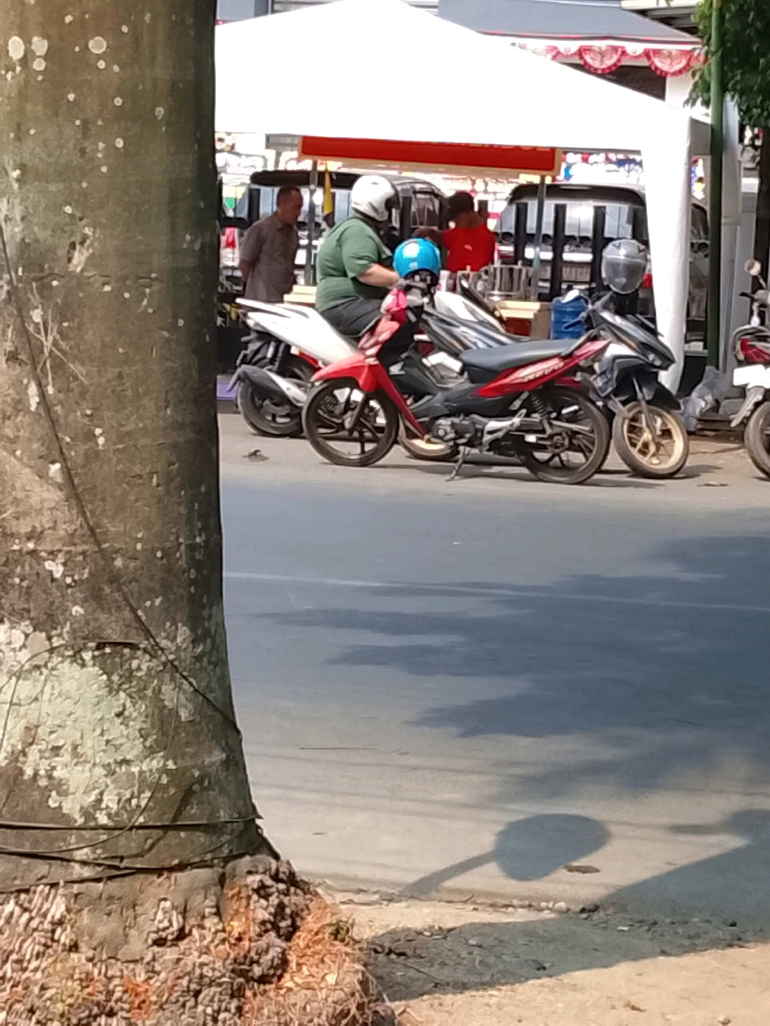 The image depicts a street scene in Slawi, Indonesia, focusing on several motorcycles parked or being operated. In the foreground, the rough bark of a large tree trunk partially obscures the view on the left. Several motorcycles are visible, with a prominent red and black motorcycle in the center, equipped with a silver helmet on its handlebars and a blue helmet on the seat. To the right of this motorcycle, another motorcycle with a black seat and silver accents is parked. Further to the right, another motorcycle with a black and white body is partially visible. In the background, under a white tent structure with red trim, a man in a green shirt and helmet is seated on a motorcycle. Another man in a grey shirt is standing to the left of the tent. The ground is paved with asphalt, and shadows are cast by the surrounding objects, indicating a sunny day.