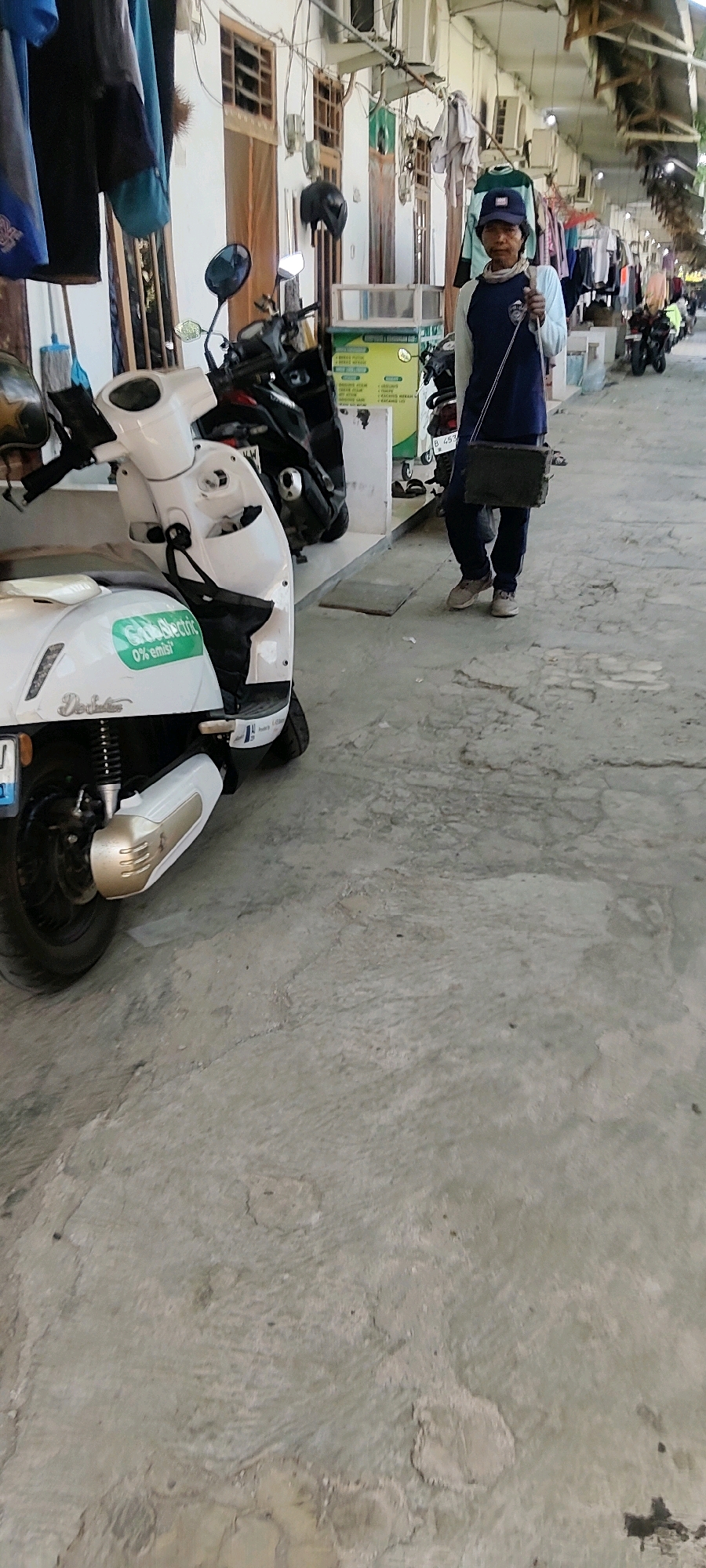 In Mekarmukti, Indonesia, a person stands on a concrete walkway in front of several shops. The person, wearing a blue baseball cap and a long-sleeved shirt, is carrying a dark rectangular object. To the left of the person is a white electric scooter with a "GrabElectric 0% emisi" sticker on its side. Behind the scooter, other motorcycles and scooters are parked. The shops appear to be open, with clothes hanging outside some of them. The overall scene suggests a commercial area with transportation and retail activity.