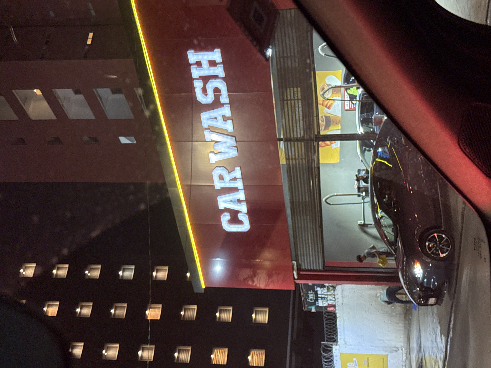 The image, taken at night from inside a vehicle, shows a vibrant "CAR WASH" facility. The main subject is a red building featuring large, illuminated white letters spelling "CAR WASH," with a bright yellow vertical light strip running alongside. Reflections of a distant building's lit windows are visible on the car wash facade.
Inside the wash bay, a dark-colored car is being cleaned by a person in a light shirt using a yellow cloth. Another person is seen crouched, attending to the front of a different car at the bottom right. Yellow posters, possibly advertisements for drinks, are visible within the bay. The ground is wet, typical of a car wash operation.
To the left, a tall, dark building with numerous evenly spaced, illuminated square windows stands in the background, suggesting an apartment or office structure. The foreground on the right is occupied by the dark red interior of the photographer's vehicle, including an A-pillar and a speaker grille. The scene depicts an active car wash in an urban setting during nighttime. A wire fence is partially visible in the lower left.