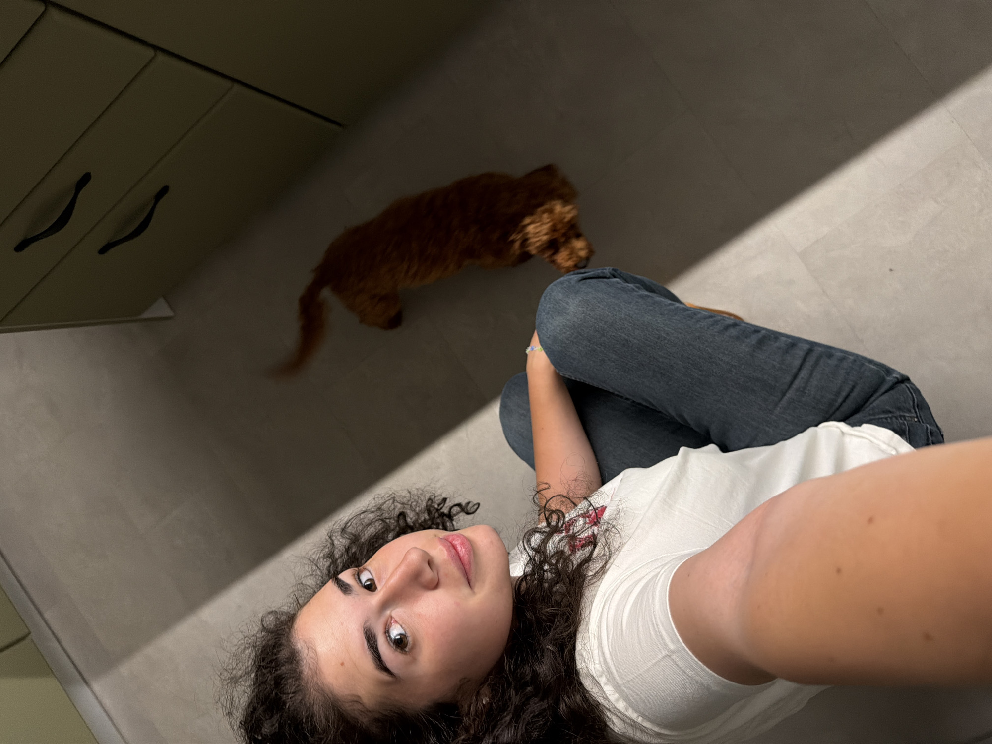 The image captures a selfie taken from a high angle, looking down on a young woman sitting on a light-colored tiled floor. She has dark, curly hair and is wearing a white t-shirt and blue jeans. To her right, a small, reddish-brown dog is visible in the background, moving away from the camera. The room appears to be a kitchen, indicated by the presence of light green cabinets with dark handles on the right side of the frame. A strong diagonal shadow cuts across the floor, suggesting a light source from the left. The woman is looking directly at the camera with a slight smile. The overall scene suggests a casual, indoor setting. There is no indication of location within Sofia, Bulgaria.
