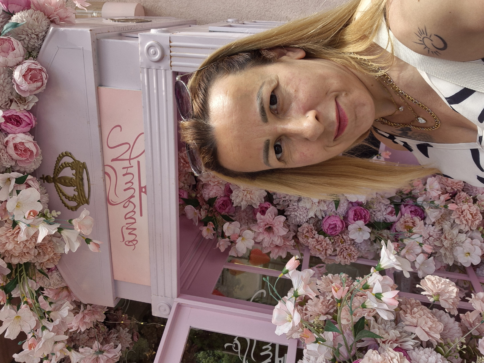 A woman with blonde hair and sunglasses on her head is taking a selfie in front of a pink decorative structure covered in artificial flowers. The structure has a crown emblem and text in cursive script. The woman is wearing a white and black patterned top and several gold necklaces. She has a tattoo of a crescent moon with a sun on her left shoulder. The background consists of more artificial flowers in shades of pink and white, and a light-colored wall. The location is not specified.