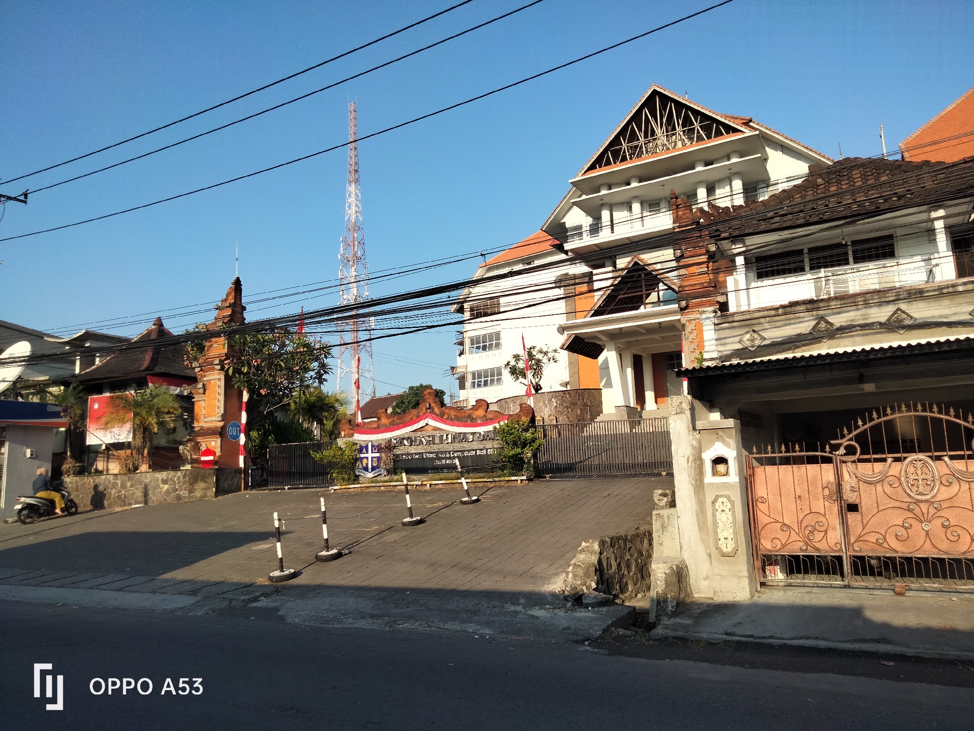 The image captures a street scene in Denpasar, Bali, Indonesia, bathed in clear, bright sunlight. The primary focus is a multi-story building with a distinctive angular roof structure, situated behind a traditional Balinese stone wall and an ornate, rusted metal gate. To the left of this building, a smaller, older structure with a reddish-brown tiled roof and intricate stone carvings is visible, possibly a temple or a traditional Balinese gate. A tall, red and white telecommunications tower stands prominently in the background against a clear blue sky. Power lines crisscross the frame, casting shadows. In the foreground, a paved driveway leads to the main building, marked by a series of black and white bollards. A person is riding a scooter on the left side of the driveway, casting a shadow on the pavement. A sign with red and white banners, featuring text and a crest, is affixed to the front of the property. The overall impression is one of a sunny day in an urban setting, with a blend of modern and traditional architecture.