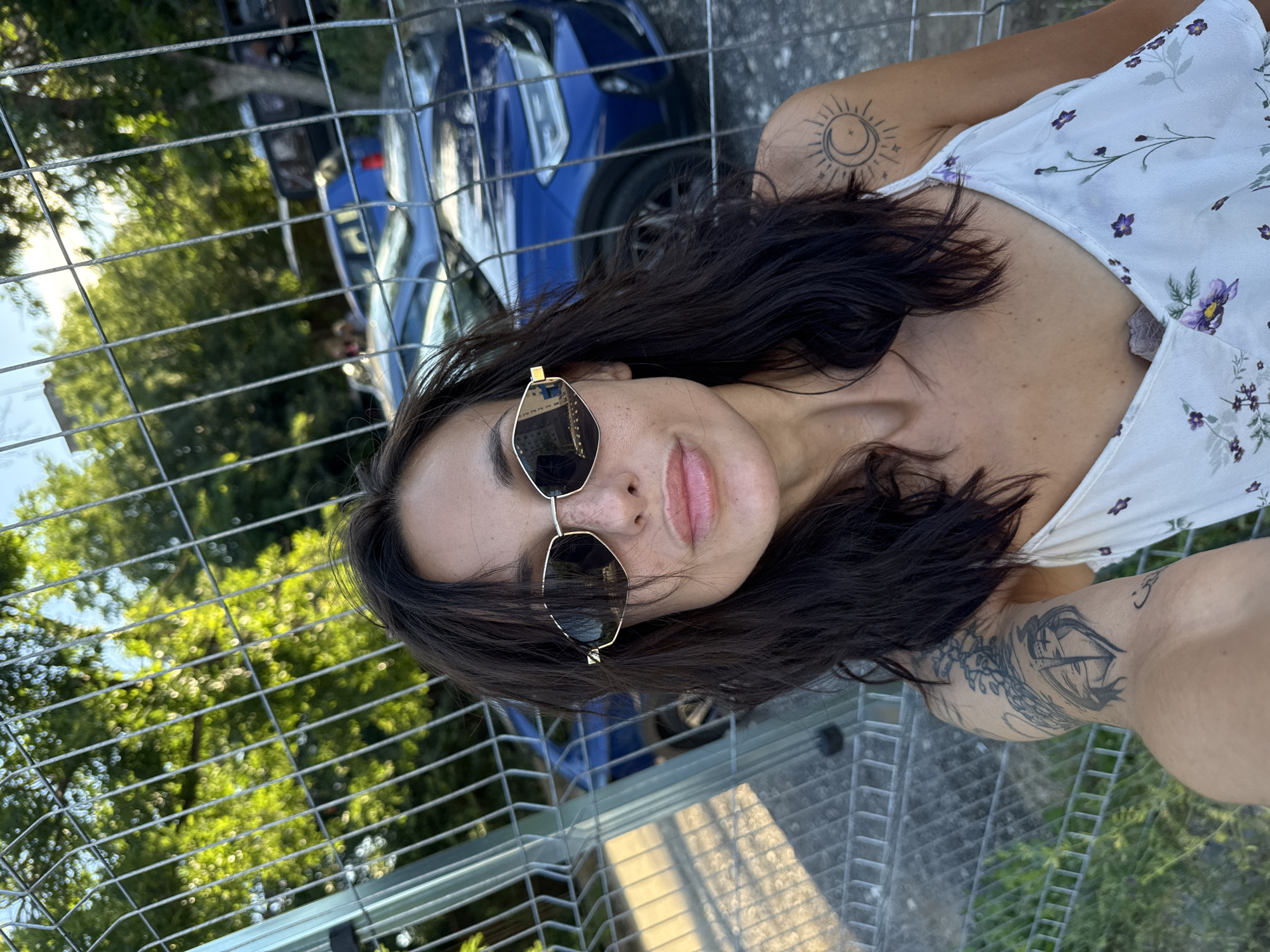 A young woman with dark hair and sunglasses is taking a selfie in front of a chain-link fence. Behind her, a blue car is parked, and trees are visible in the background. The woman is wearing a white floral top. There are tattoos on her arm and shoulder. The scene appears to be outdoors, possibly in a parking lot or a residential area, in the city of Pleven, Bulgaria.