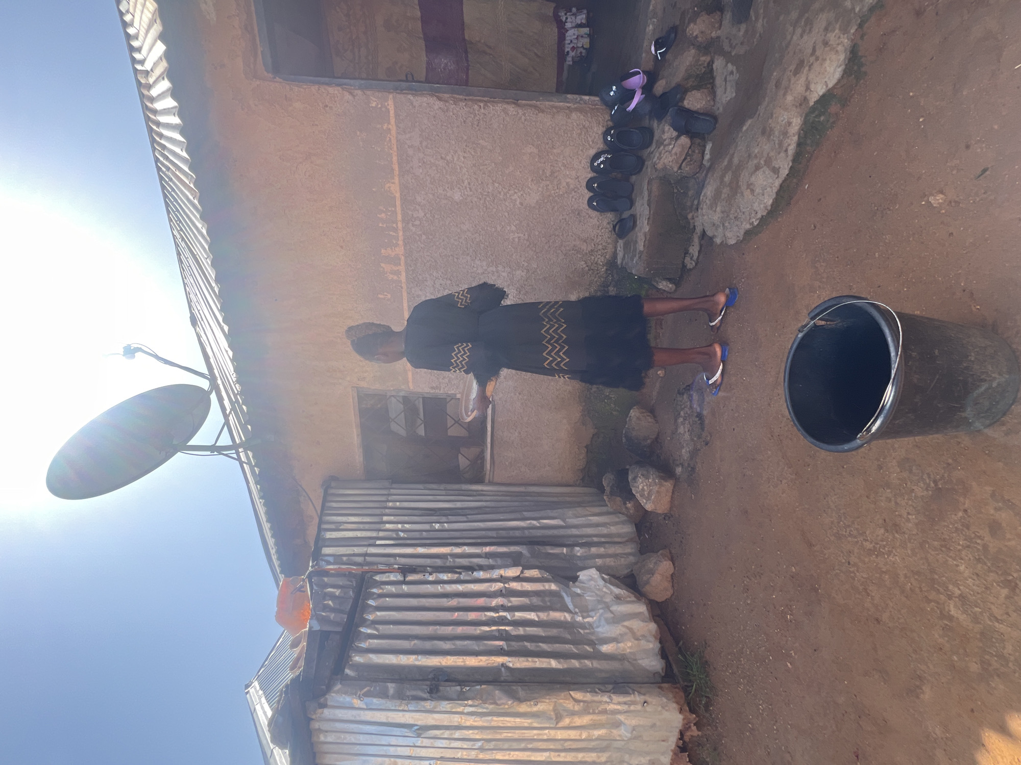 In an outdoor scene in the city of Jos, Nigeria, a young person stands in front of a stucco-walled building with a corrugated metal roof. The person is wearing a dark dress with a patterned design and appears to be holding a plate. Several pairs of sandals are scattered on the ground near a rocky ledge. To the left of the building, a structure made of corrugated metal sheets is visible. A large black bucket sits in the foreground on the dirt ground. The sky is bright blue with a strong sun, casting lens flare across the image.