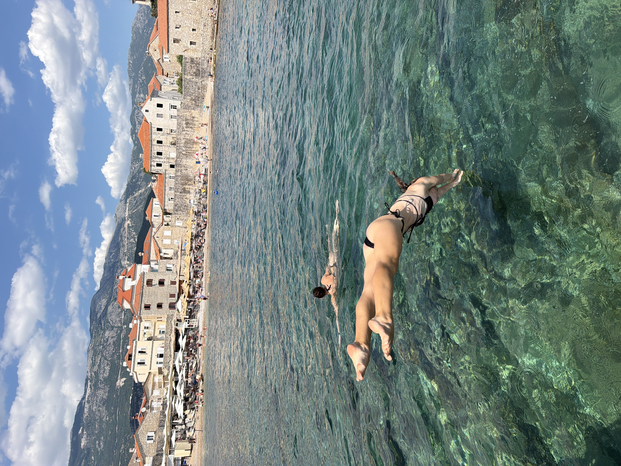 The image captures a sunny day at a beach in Budva, Montenegro. In the foreground, a person in a black bikini is captured mid-dive into the clear, turquoise water. Their body is in a streamlined position, with legs extended and feet pointed. Further in the water, another person is swimming.
The background shows a crowded beach lined with buildings that have red-tiled roofs. The architecture suggests a historic European coastal town. Beyond the buildings, a majestic mountain range rises, covered in lush green vegetation. The sky is a vibrant blue, dotted with fluffy white clouds, indicating fair weather. The sun is high, casting bright light on the scene, suggesting it is midday.
The overall atmosphere is one of leisure and recreation, with people enjoying the beach and the sea on a beautiful summer day. The clear water offers visibility of the seabed, adding to the picturesque quality of the scene. No visible text is present in the image.