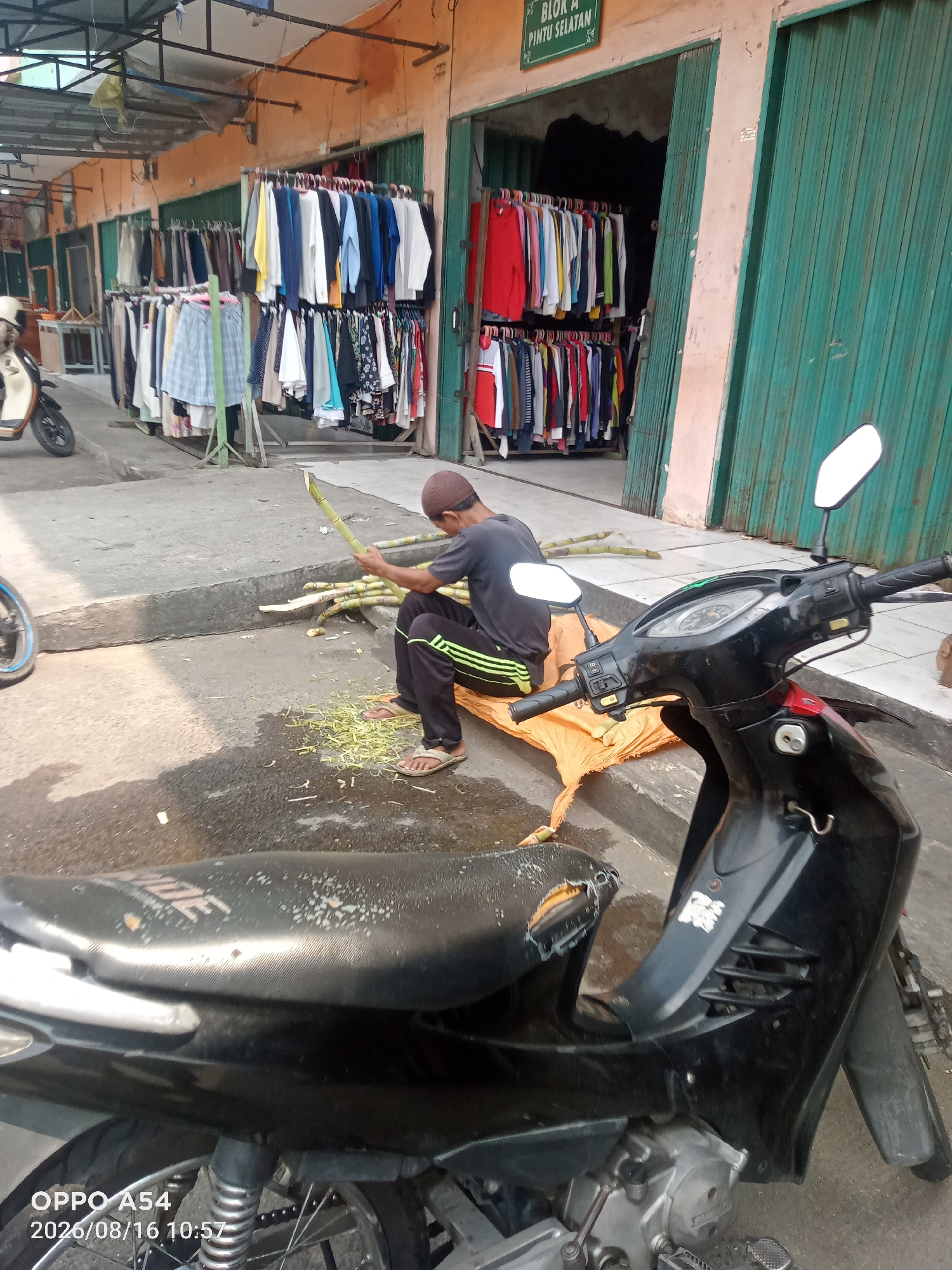 The image depicts an outdoor scene, likely in Jambi, Indonesia, near a row of market stalls. In the foreground, a black motorcycle is parked. To the left of the motorcycle, a young man wearing a brown cap and a dark t-shirt with green accents on his pants is seated on an orange tarp. He is in the process of cutting sugarcane stalks, with several pieces already cut and scattered around him. Behind the man and the motorcycle, several market stalls are visible, displaying a variety of clothing items on racks. The stalls have orange and green colored fronts. The ground appears to be paved with concrete and asphalt. A sign with green lettering is visible above one of the stalls. The overall atmosphere suggests a busy market environment, possibly during daylight hours.
