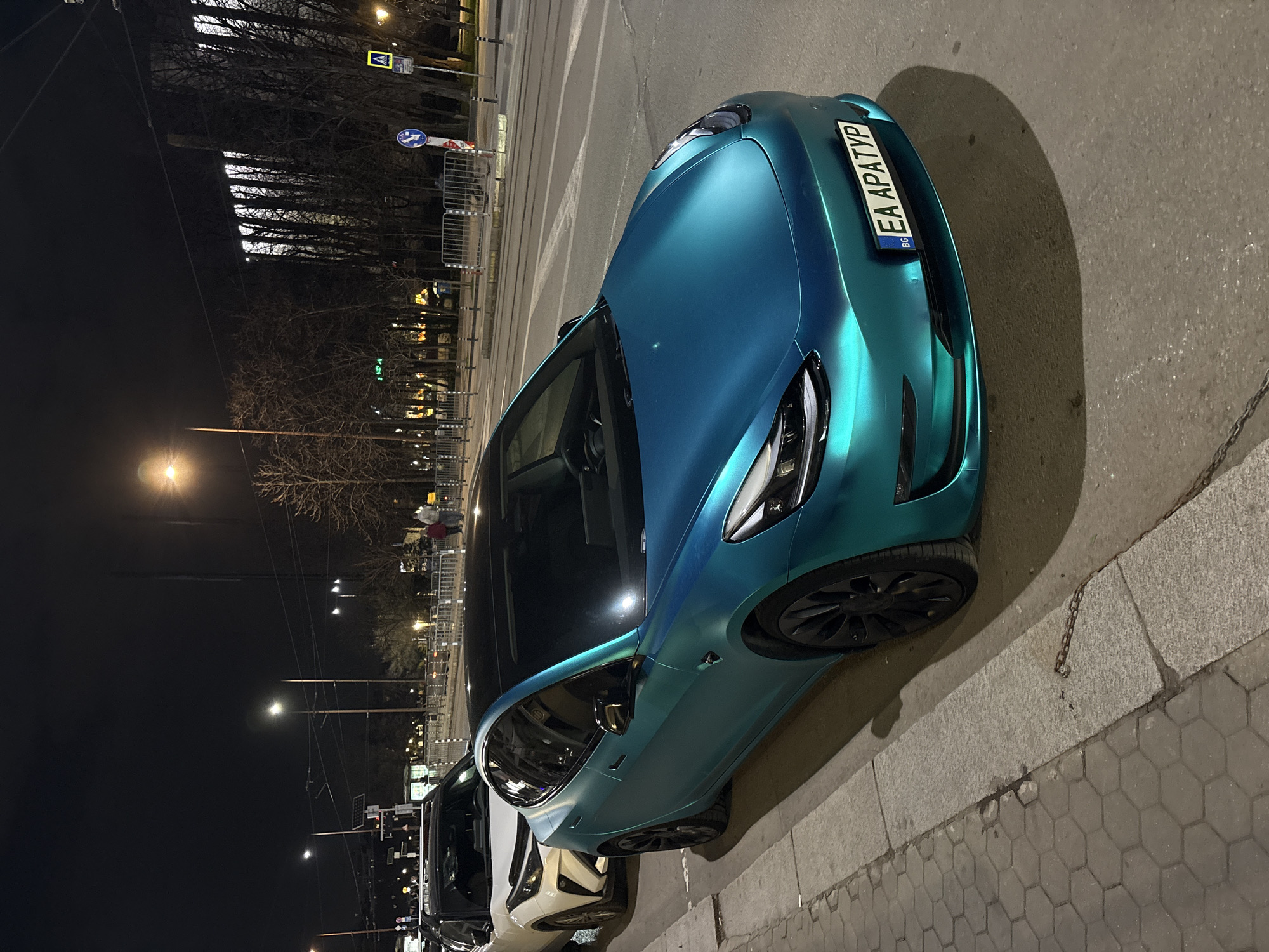 The image depicts a nighttime street scene in Sofia, Bulgaria, specifically identifiable by the distant architectural profile of the National Palace of Culture (NDK) in the background. In the foreground, a teal/blue-green matte-wrapped Tesla Model 3 is parked parallel to a concrete curb, facing slightly right. Its front license plate displays "BG EA APATYP". Behind the Tesla, a white Toyota C-HR is also parked on the street. The sidewalk features hexagonal paving stones. A short section of a metal chain lies on the curb next to the Tesla.
The asphalt road surface in front of the cars shows faint white road markings, and tram tracks are visible further into the street. The environment is illuminated by streetlights, casting distinct shadows from the parked vehicles. Bare trees line the street, and a pedestrian crossing sign (yellow square with black pedestrian symbol) and a blue circular traffic sign are visible on the right side of the road. Metal crowd control barriers are positioned along the sidewalk and a median strip. Two indistinct human figures are discernible walking on a distant sidewalk in the upper right quadrant of the image. The overall lighting suggests artificial illumination from streetlights and building lights.