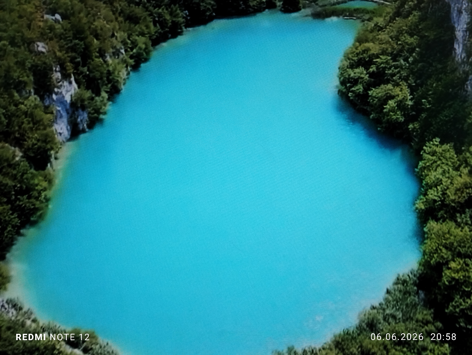 An aerial or elevated view captures a naturally shaped body of water, likely a lake or lagoon, displaying a vibrant turquoise color. The water is bordered by dense green vegetation. The left bank features a prominent light-colored rocky cliff face, integrated with trees and foliage. The right and bottom shores are heavily forested with dark green trees. A narrower inlet or connecting waterway, also tree-lined, is visible at the top of the scene. No individuals, animals, or artificial structures are discernible within the visible landscape. Digital overlays indicate "REDMI NOTE 12" in the bottom left corner and "06.06.2026 20:58" in the bottom right corner. A fine grid pattern is present across the image, indicative of a photograph of a digital screen. This scene is located in the city of Burgas, Bulgaria.