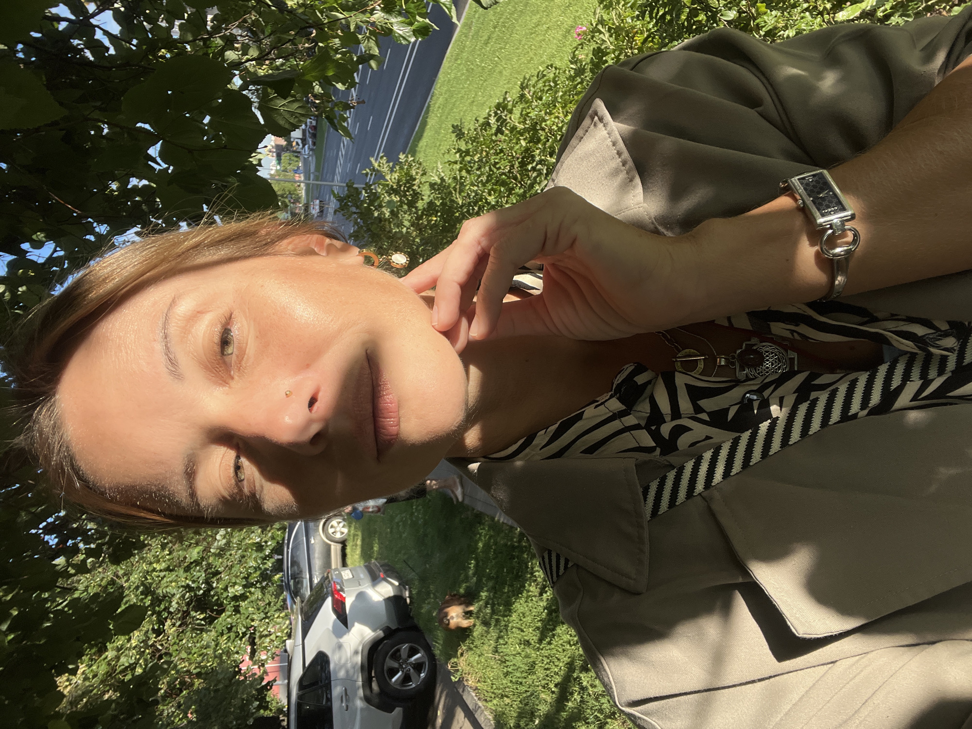 A woman is taking a selfie outdoors in Moscow, Russia. She is outdoors, with trees and a street visible in the background. The woman has her hand on her chin, and she is wearing a light-colored trench coat over a black and white patterned shirt. She is also wearing a silver watch and a necklace with several pendants. A small brown dog is visible on the grass in the background, near two parked cars. The sun is shining, casting shadows on the woman and the surrounding environment.