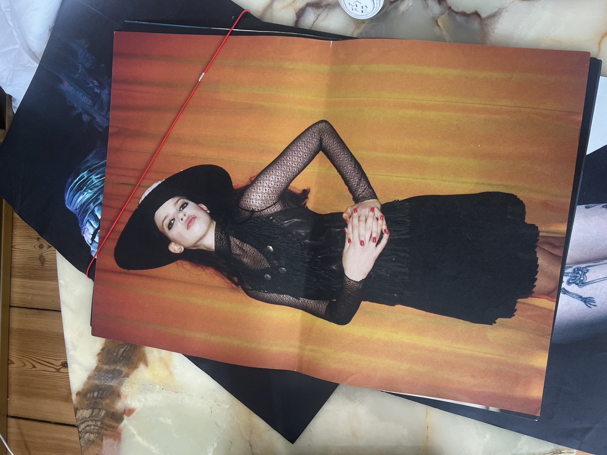 The image shows a magazine open to a page featuring a woman lying down in a black dress and a wide-brimmed black hat against a warm-toned orange and yellow background. Her arms are positioned above her, with her hands near her face. She has red nail polish. The magazine is lying on a marble-patterned surface. A red object (possibly a string or wire) stretches across the magazine. Other objects are partially visible around the magazine, including another image with a blue and black pattern and a portion of a leg with a tattoo. A partially visible object on the right side might be a white container. The location, Berlin, Germany, cannot be determined from the image content.