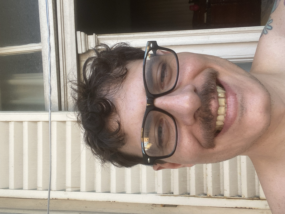 A person smiles with visible teeth, wearing black-rimmed glasses and sporting a dark mustache. The individual has curly dark hair and a fair complexion. The background features a white window frame and what appears to be an exterior wall with vertical slats. A glimpse of an interior room is visible through the window. The picture is taken at an angle, tilting the subject to the side. A tattoo can be seen on the person's right shoulder.
