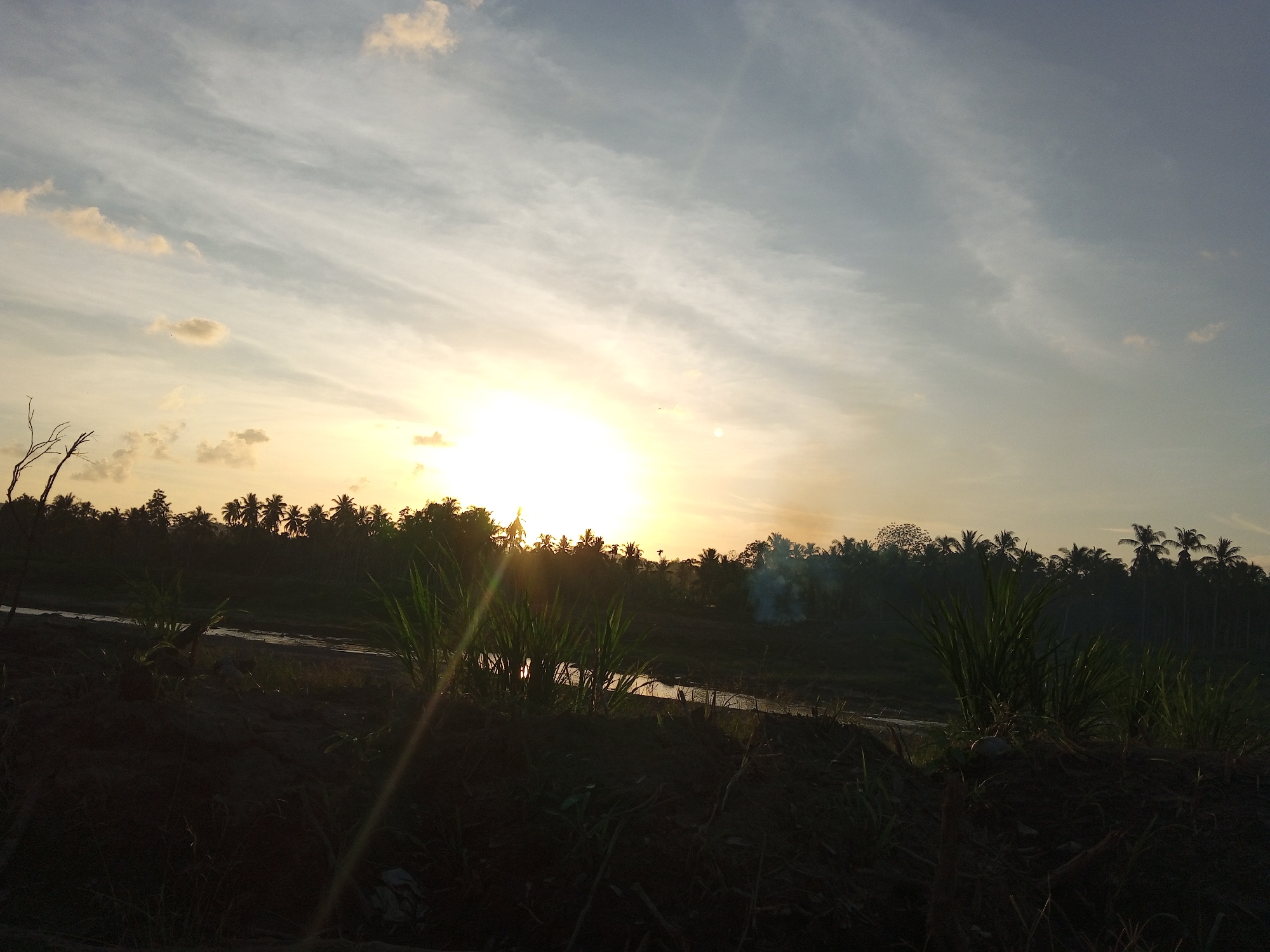 The image depicts a landscape at sunset in Kubu, Indonesia. In the foreground, there is dark vegetation on a raised bank. Behind this, a narrow river or stream reflects the bright light of the setting sun. The background consists of a silhouette of trees, primarily coconut palms, against the bright sky. There is a small plume of smoke rising from the trees on the right side of the image. The sky is a mix of pale blue and golden hues, with streaky clouds.