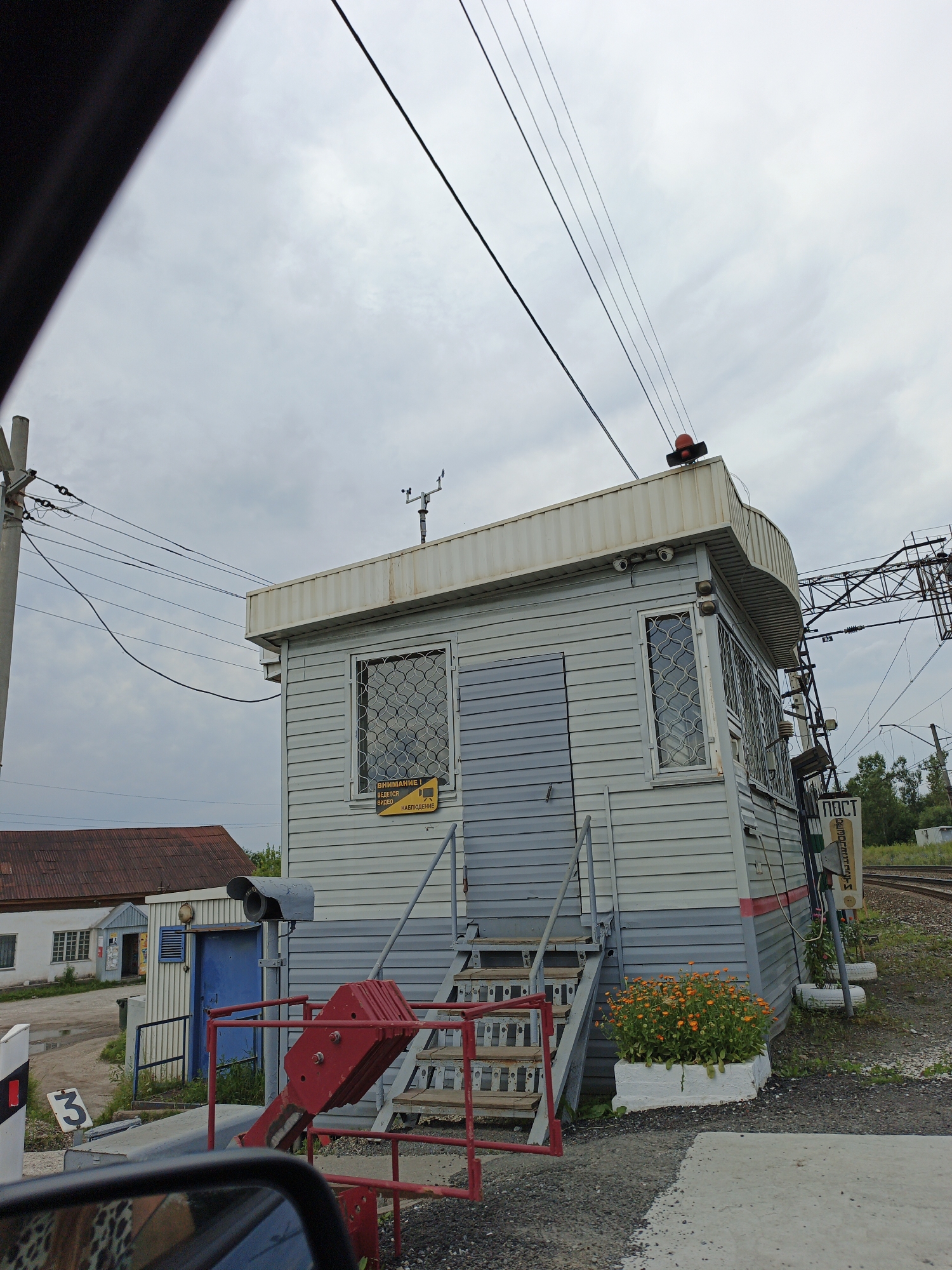 The image depicts a railway crossing guard house in Medvedyovka, Russia, under an overcast sky. The light gray, two-story structure has horizontal siding and a white, corrugated metal roof. A red signal light is positioned on the top of the building. Two windows with patterned security bars are visible on the front, and a gray door is centrally located. A metal staircase with wooden treads leads up to the entrance.
To the left of the building, a smaller white structure with a blue door and windows is visible. In front of the guard house, there is a red metal barrier and a red lever mechanism. A grey security camera is mounted on a pole to the left of the blue door. A planter with orange flowers sits at the base of the stairs on the right side of the guard house.
A sign with Cyrillic text, reading "ПОСТ" (Post) is vertically mounted on a pole near the tracks to the right of the guard house. The railway tracks are visible in the background, curving to the right. Several utility poles and overhead wires are present in the scene. A sign with the number "3" in a black circle is visible in the lower left corner of the image, next to a white and red striped marker. The foreground shows a portion of a car's side mirror.