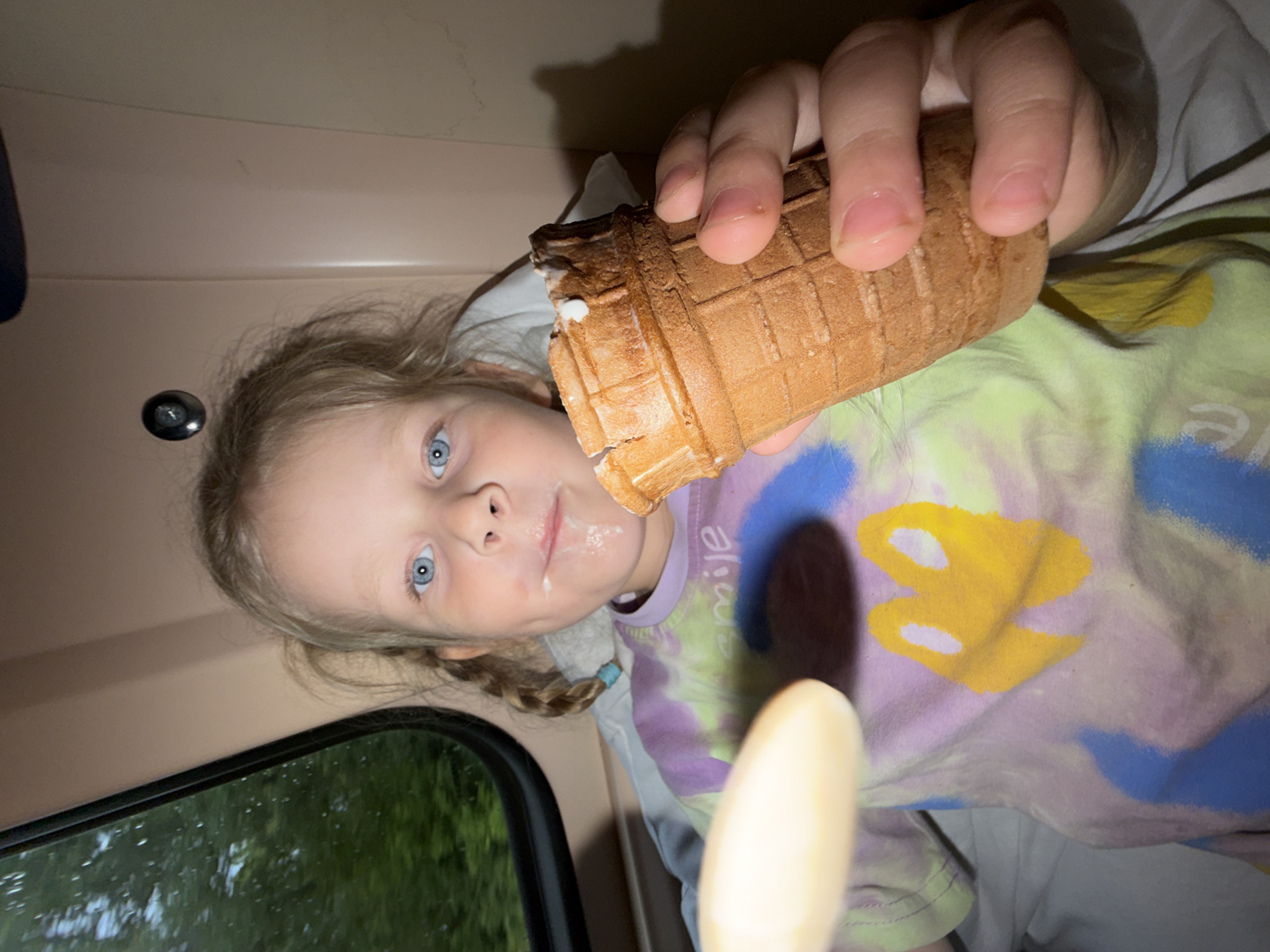 A young girl with blonde hair in pigtails is holding an ice cream cone and a spoon. She has ice cream on her face. She is wearing a tie-dye shirt with a yellow graphic and the word "smile". The girl is seated inside a vehicle, possibly a train, as indicated by the window showing trees and greenery outside. The interior of the vehicle appears to be beige and brown.