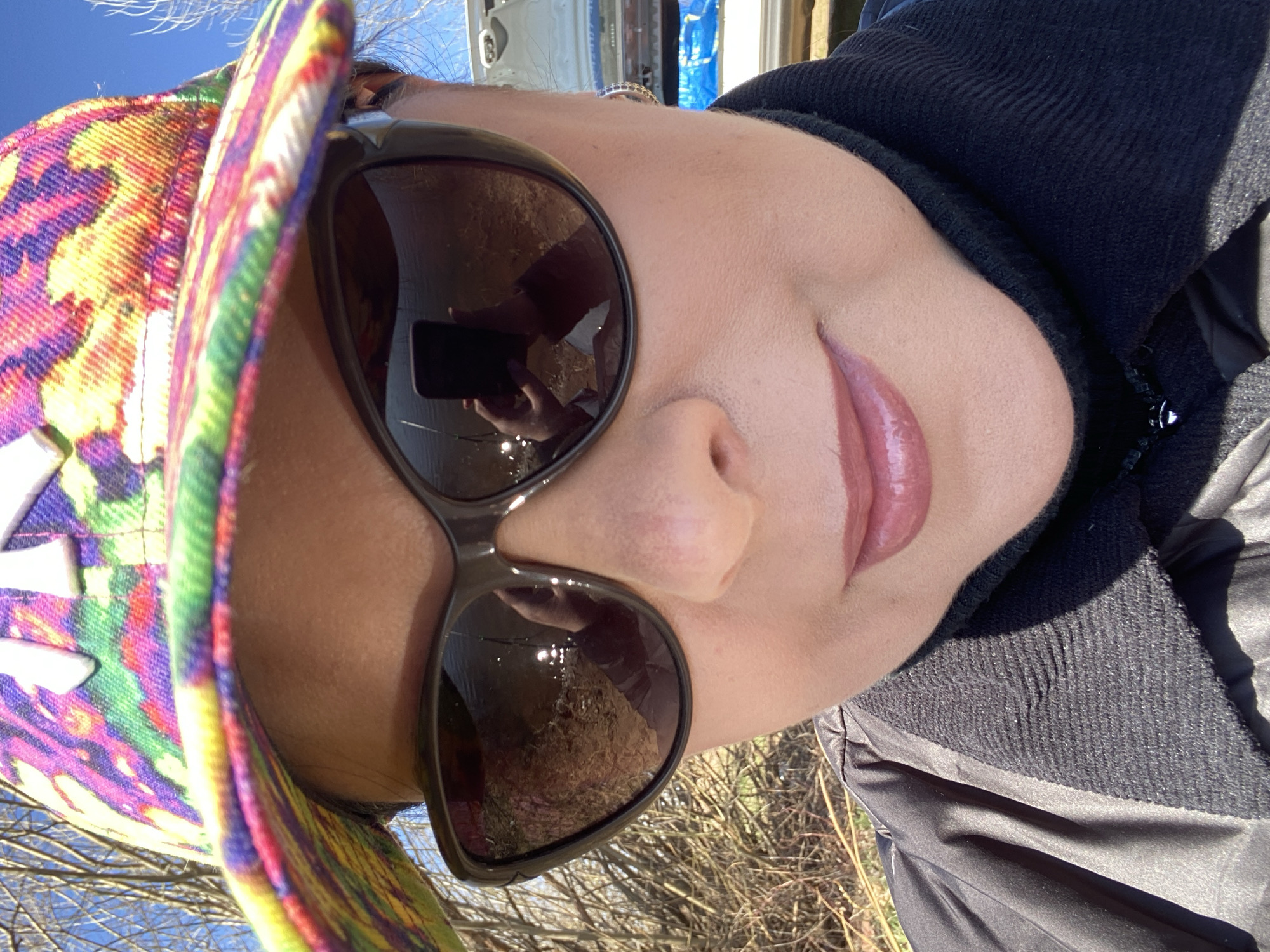 A close-up, tilted image shows a person with light skin wearing sunglasses and a multi-colored hat. The person has full lips and is wearing a black jacket. The reflection in the sunglasses suggests an outdoor environment with trees or foliage. The background also shows a sliver of blue sky and what appears to be part of a vehicle. There is no information present in this image to definitively place the location in the city of Ольховка, Russia.