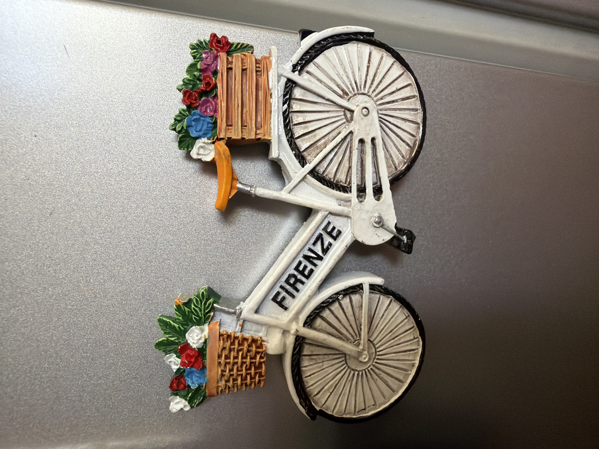 A decorative white bicycle magnet with two baskets of flowers is attached to a silver metallic surface, likely a refrigerator. The bicycle has a prominent basket on the front, filled with red, white, and blue flowers and green leaves. The word "FIRENZE" is emblazoned on the frame of the bicycle in black lettering. A second, smaller basket is attached to the rear of the bicycle, also containing flowers and greenery, primarily in shades of pink, red, and blue. The bicycle has orange handlebars and a seat. The surface it is attached to has a textured, metallic finish. The context of Pazardzhik, Bulgaria, is not evident from the image itself.