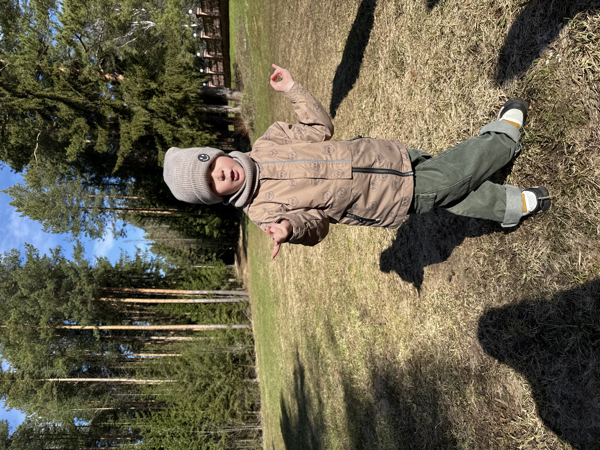 A young child lies on a grassy field, partially shaded by trees, on a sunny day. The child is wearing a beige patterned jacket, green pants, a grey beanie with a smiley face, and black and white shoes. The child is facing the viewer and gesturing with both hands. The ground is covered in dry grass, and tall trees can be seen in the background. The exact location in the city of Адам, Russia cannot be determined from the visual evidence.