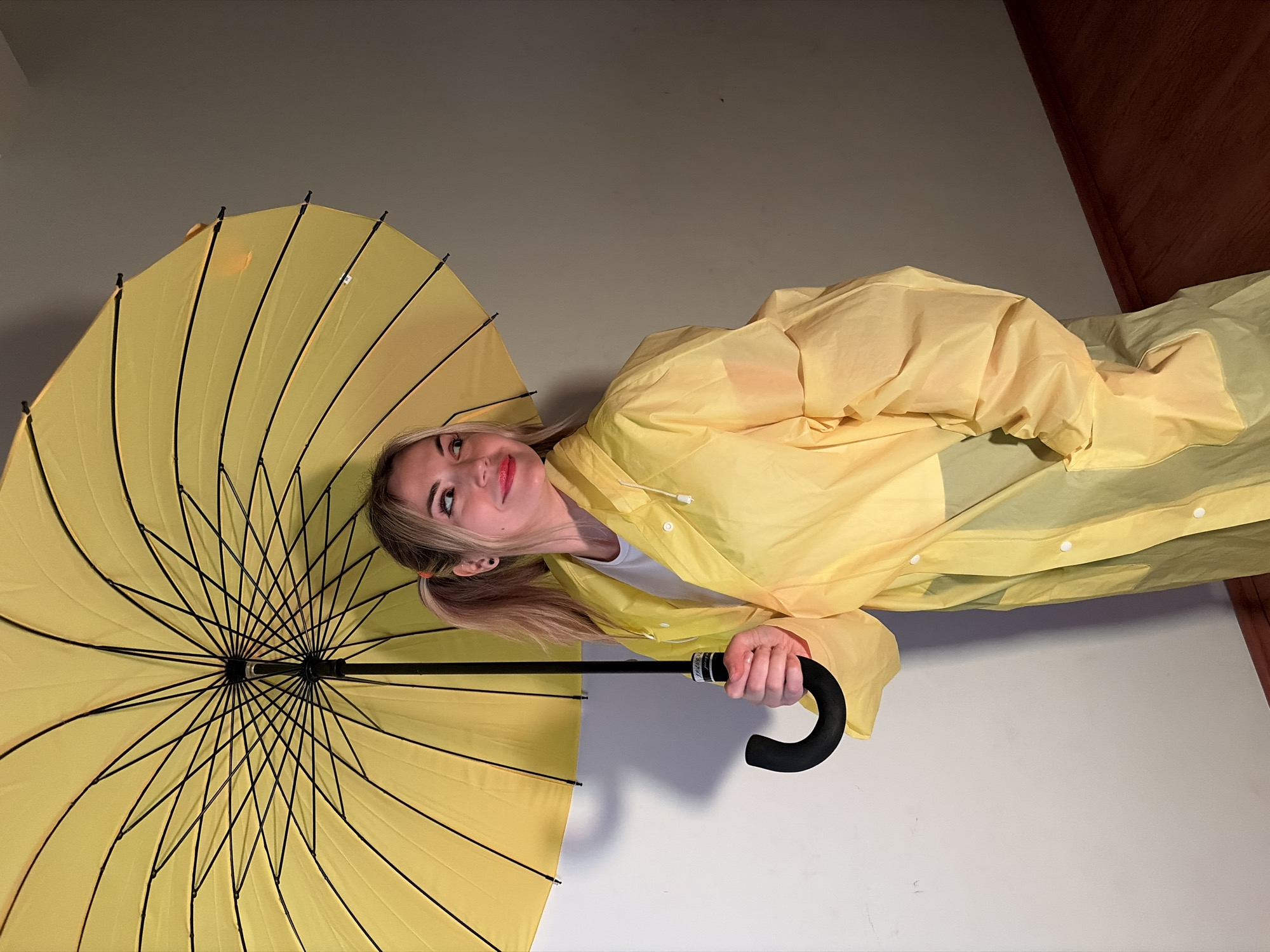 A young woman is depicted in a portrait-style shot, rotated 90 degrees clockwise from its original vertical orientation. She has fair skin, blonde hair styled in pigtails, and is wearing makeup including red lipstick. She is dressed in a bright yellow raincoat over a white t-shirt and holds a large, open yellow umbrella with a black handle and spokes.
The woman is posing, looking upwards and slightly to her right, with a soft, pleasant smile suggesting contentment or playfulness. Her left hand firmly grips the curved black handle of the umbrella, which is prominently featured on the left side of the frame, partially obscuring her head. The umbrella's intricate black metal framework is clearly visible against its yellow canopy.
The background consists of plain, neutral-toned walls – a light grey on the left and a darker, possibly brown or dark grey, section on the right, suggesting an indoor or studio setting. A draped surface, perhaps a bed or furniture, is visible in the bottom right corner of the image. The lighting appears soft and even, characteristic of an artificial indoor setup. There are no direct visual cues to the weather, but the raincoat and umbrella imply a theme of rain. Indistinct text, possibly a brand name, is visible on a label attached to the umbrella handle. The image was taken in Budva, Montenegro.