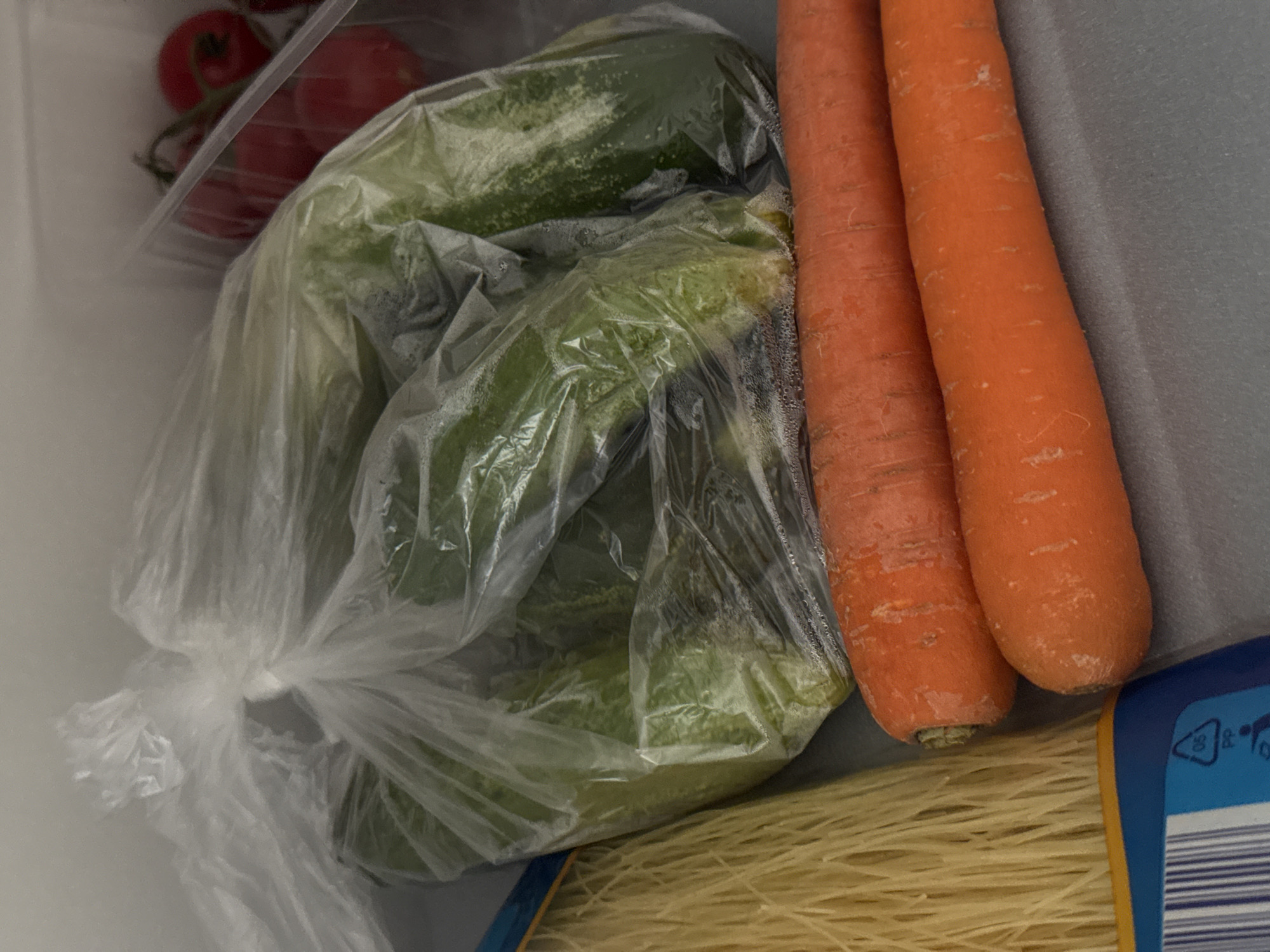 The image shows a close-up view of food items stored inside a refrigerator. In the foreground, two large, bright orange carrots are positioned horizontally. Behind the carrots, to the left, is a bundle of thin, pale yellow pasta. Dominating the center and upper portion of the image is a clear plastic bag containing several green vegetables, which appear to be cucumbers, with visible condensation on the inside of the bag. To the upper right, a clear plastic container holds a cluster of ripe red cherry tomatoes on the vine. The background is a plain, light gray surface, consistent with the interior of a refrigerator. There are no people or animals present in the image. The lighting suggests a typical indoor refrigerator environment. The scene depicts a collection of fresh produce ready for meal preparation. There is no information available to determine the location in the city of Bratislava, Slovakia.