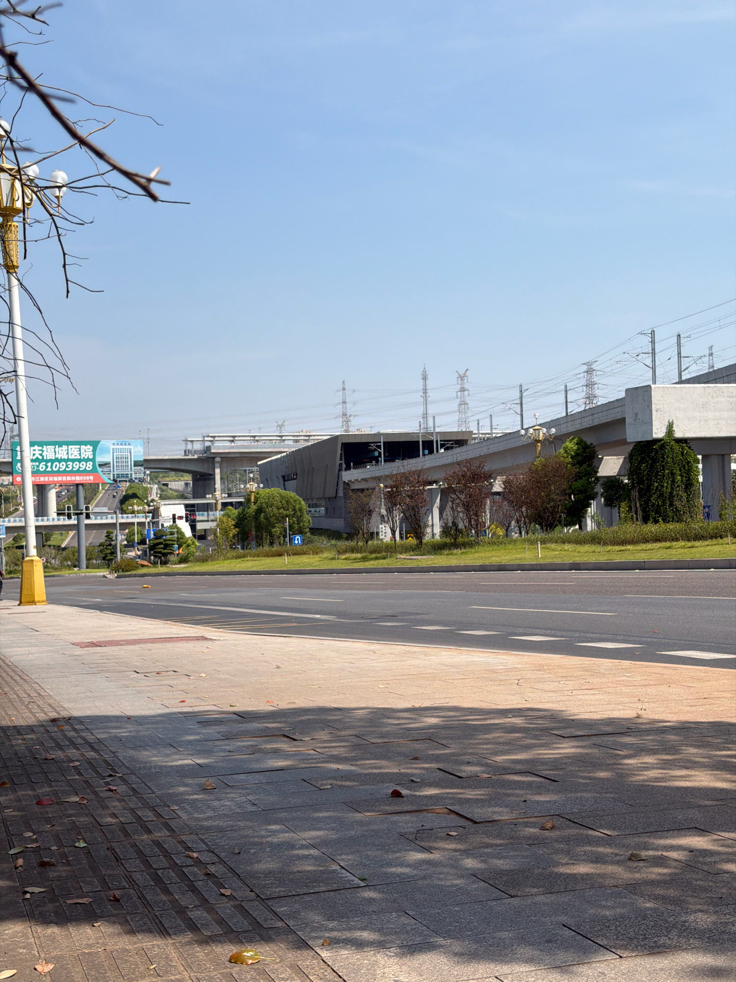 The image shows a street scene in the Jiangjin District of Chongqing, China, on a clear, sunny day. A wide road occupies the lower right portion of the frame, with a pedestrian sidewalk on the left. Overhead, an elevated structure, likely a railway or highway overpass, spans across the scene, supported by concrete pillars. Electrical transmission towers and power lines are visible in the background, indicating an urban or suburban environment.
To the left of the road, a billboard is prominently displayed on a large pillar. The billboard features Chinese text and images, including what appears to be a hospital building and contact information, with the characters "庆福城医院" (Qingfu City Hospital) and a phone number "23-61093998" visible. A traffic light is also present near the billboard.
A streetlamp with a decorative yellow base and white globes stands near the sidewalk. The sidewalk itself is paved with square tiles, some of which appear worn or slightly dislodged. Fallen leaves are scattered across the sidewalk and the adjacent grass verge.
In the mid-ground, a section of the elevated structure passes over a well-maintained grassy area with several small trees. The elevated structure is concrete and appears to be part of a modern transportation network. The sky is a clear blue with no visible clouds. The overall impression is one of a developed urban area with ongoing infrastructure. There are no people clearly visible in the scene.