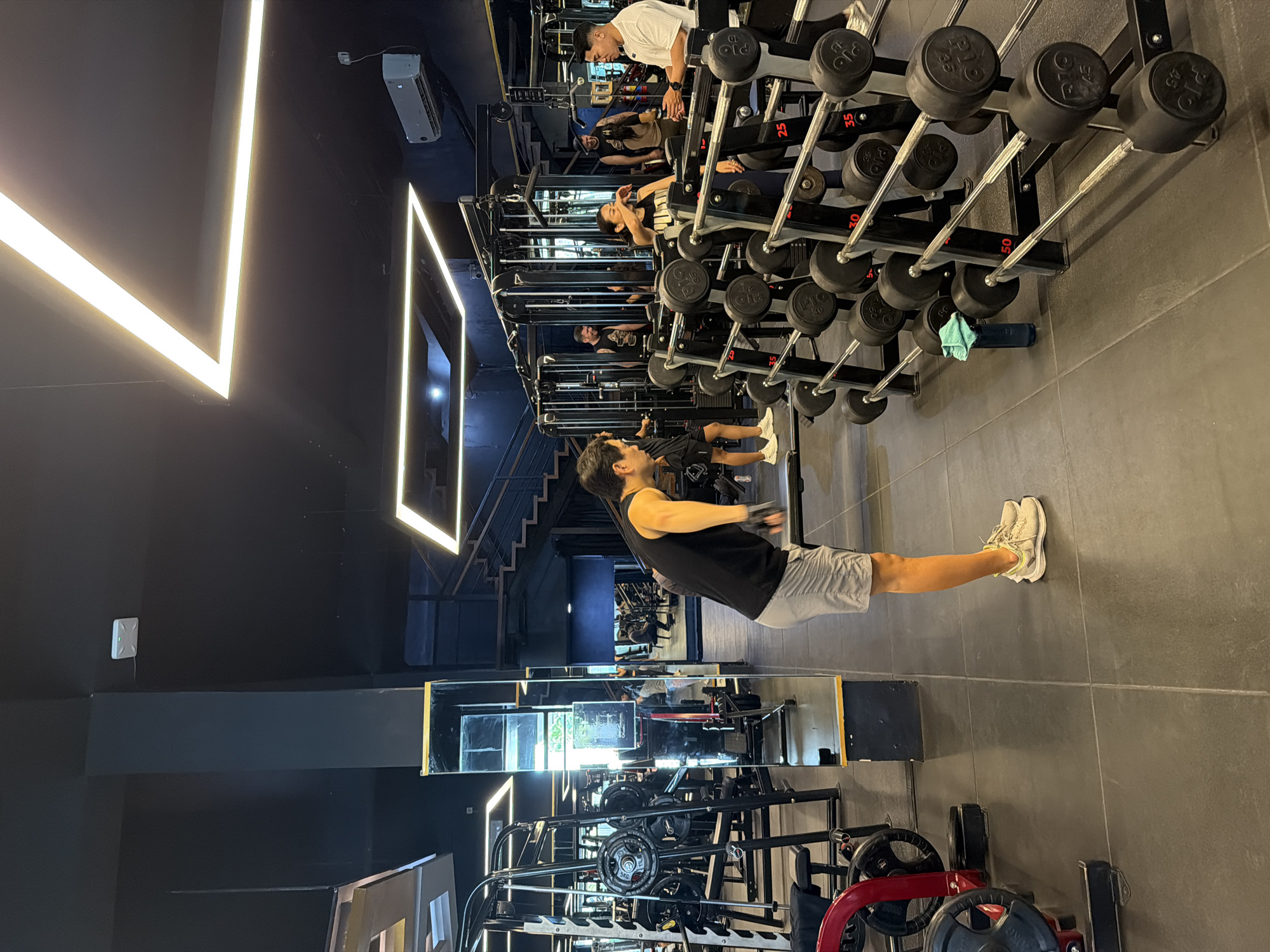 This image captures the interior of a gym located in Denpasar, Indonesia. A man in a black tank top and gray shorts is performing a stretch on the gray tiled floor. To the right of the man are racks of barbells. In the background, other gym-goers can be seen using weight machines. The gym has black walls and geometric, neon lighting fixtures are visible on the ceiling. A mirror runs along the bottom of the wall, reflecting exercise equipment.