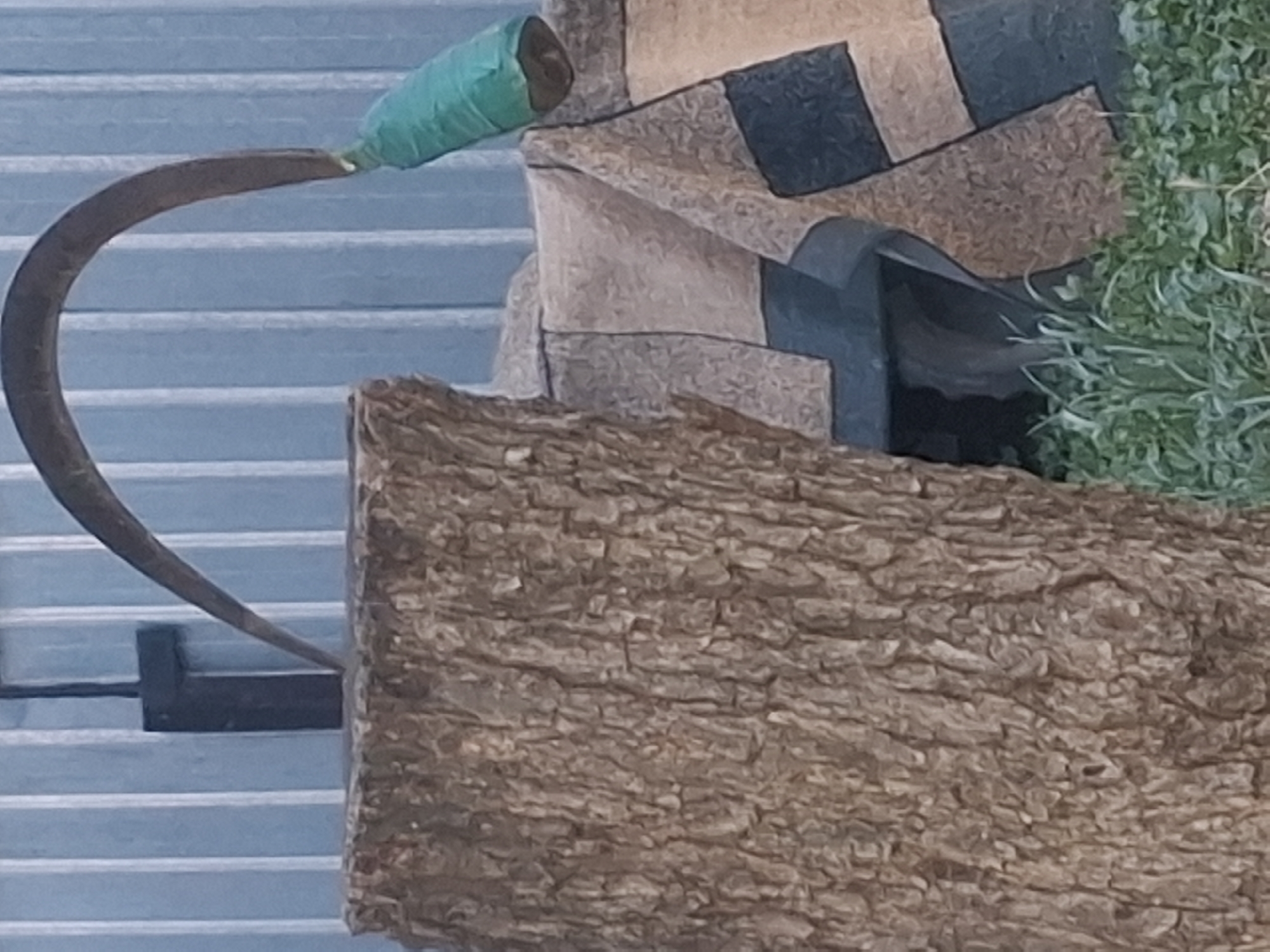 This is a close-up, medium shot of an outdoor, decorative arrangement. The most prominent subject is a rustic, textured piece of bark, filling the lower right portion of the frame. In the upper left, a dark, curved metal object, possibly part of a sickle or scythe, extends into the frame from the left. Its handle is wrapped in bright green tape. Behind the metal object and to the right of it, a patchwork of earthy tones – browns, tans, and dark blues – creates a geometric pattern. To the far right, vibrant green foliage, resembling moss or a small plant, is visible.
The background consists of horizontal light blue slats, suggesting an outdoor structure like a fence or siding. The lighting appears to be natural daylight, casting soft shadows. There are no people visible, and no specific activity is clearly depicted; the scene appears to be a static display or part of a larger artistic installation. The overall impression is one of natural and manufactured elements combined in a decorative manner. Given the context of Almaty, Kazakhstan, this could be part of a garden decoration, a public art piece, or a creative display outside a building. The image lacks any visible text.