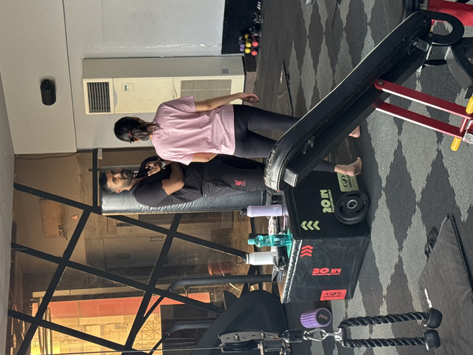 A man and woman stand near gym equipment in a fitness center. The woman wears a pink shirt and black pants, while the man is in a black shirt and pants. A black step-up box with water bottles rests next to them. Weight training equipment and black and gray patterned flooring are visible. An air conditioning unit is on the wall. The image is rotated.