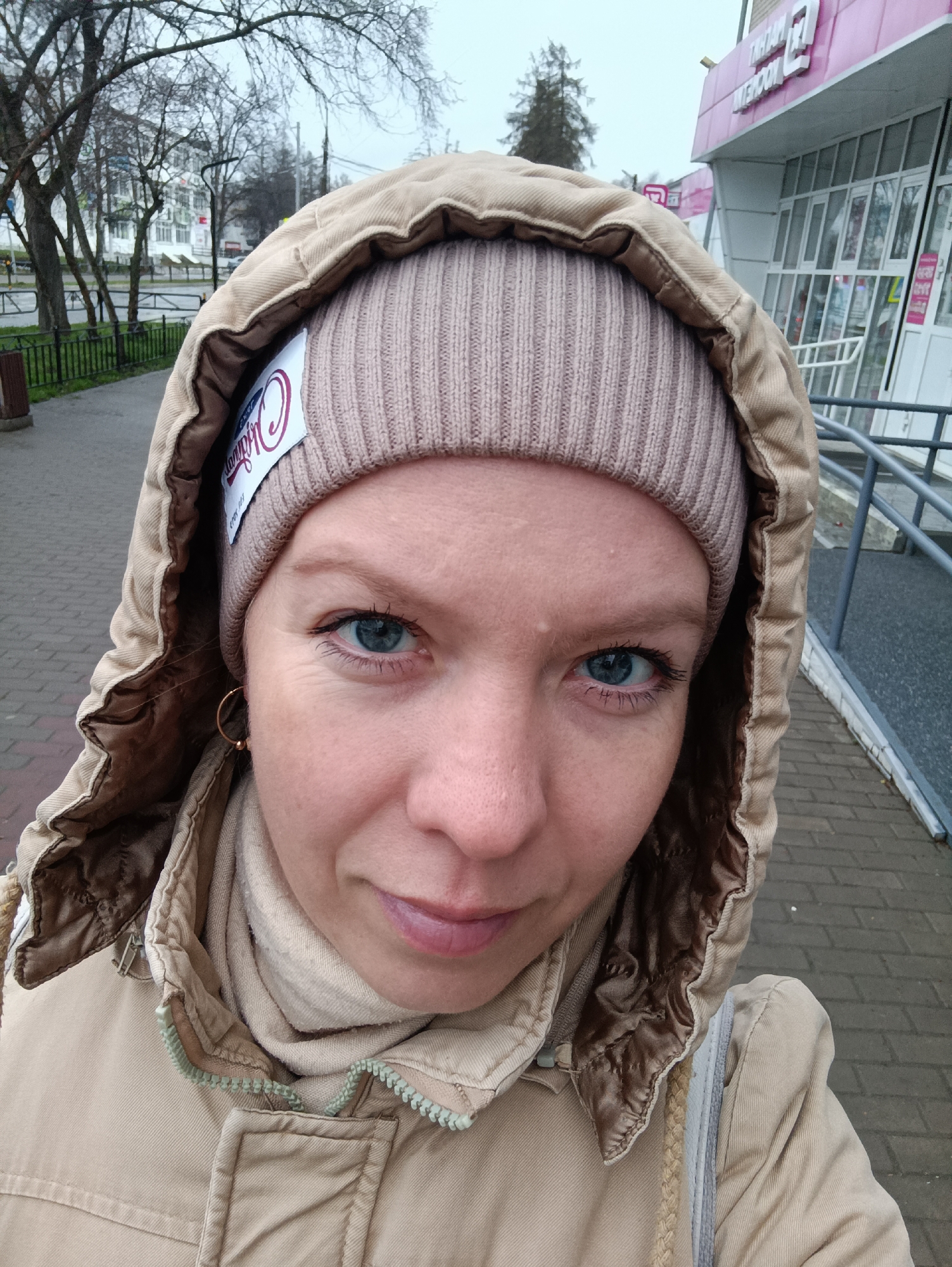 A close-up selfie shows a person with light skin, blue eyes, and wearing a beige knit hat under a hooded tan jacket. The hood is lined with a shiny brown fabric. A light beige scarf is visible around the person's neck, and a gold hoop earring is seen on the left ear. A white label with a logo is affixed to the knit hat.
The person is standing on a brick sidewalk, with a building in the background on the right. The building has a pink sign with lettering in Russian. In the background to the left, trees and other buildings are visible. The location is likely Chaikovsky, Russia based on the Russian signage.