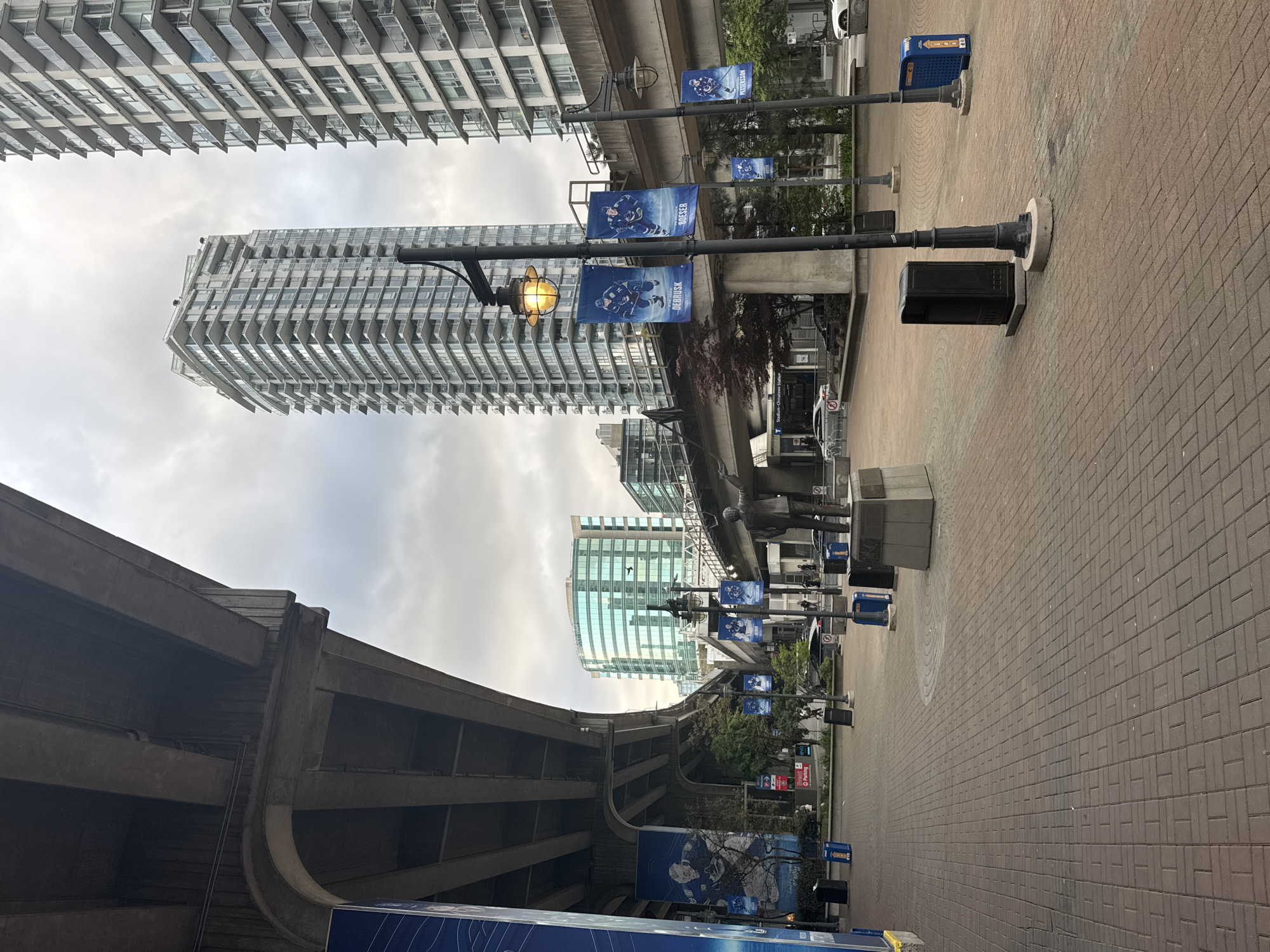 The image displays an urban street scene in Vancouver, Canada, characterized by modern architecture and an overcast sky. The photograph is oriented vertically, rotated 90 degrees clockwise.
Tall, contemporary high-rise buildings, likely residential or office, frame the scene. On the left, a prominent white skyscraper with numerous staggered balconies reaches towards the sky. Further down and to the right, another similar building is visible, and in the background, a glass-faced building reflects the sky. An elevated concrete structure, resembling a SkyTrain track, cuts across the middle of the frame, contributing to the urban infrastructure.
A wide pedestrian pathway, paved with reddish-brown interlocking bricks, extends into the distance on the right. Several black lamp posts line this path; one features an illuminated yellow lamp. Blue banners displaying hockey players in action are hung from these posts. Visible names on the banners include "PETTERSSON," "BOESER," and "DEBRUSK," suggesting an association with the Vancouver Canucks hockey team. The word "ROGERS" is also partially visible on one banner. Black trash cans and blue recycling bins are positioned along the walkway.
A crucial location cue is a sign on a pole that clearly states "Stadium-Chinatown Station," identifying a known SkyTrain station in Vancouver. Another small sign in the distance indicates "P Parking." The sky is entirely cloudy and overcast, suggesting a daytime scene without direct sunlight. No people are visible, giving the area a quiet, uninhabited atmosphere.