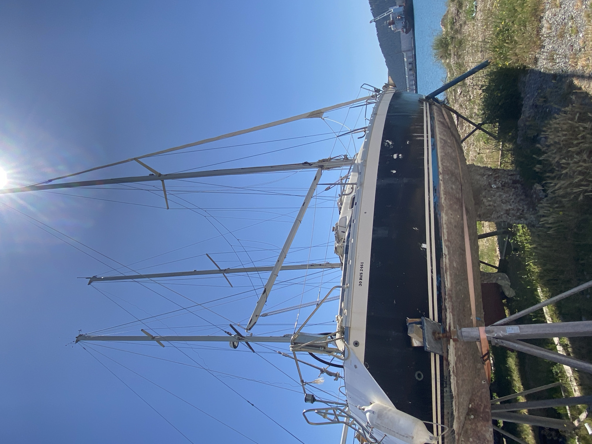 The image displays a large sailboat positioned on dry land, suggesting it is in a boatyard or dry dock. The vessel features a black hull accented by a white stripe and a white superstructure. Multiple masts with extensive rigging are prominent against a clear, bright blue sky, indicating a multi-masted sailboat. The sails are not deployed, implying the boat is either in storage or undergoing maintenance. Several white fenders hang from the boat's side.
The sailboat is securely held upright by a combination of metal stands and a wooden cradle structure. On the white section of the hull, the alphanumeric text "30 RUS 2461" is clearly visible. A strong sun flare emanates from the upper left portion of the frame, signifying a very sunny day, likely during midday or early afternoon.
The ground immediately surrounding the boat is unpaved, characterized by rocky terrain, dry grass, and scattered vegetation. In the background, a calm body of water, presumably a harbor or bay, is visible, with distant land or possibly industrial structures along the far shore. This maritime scene is located in Bar, Montenegro.