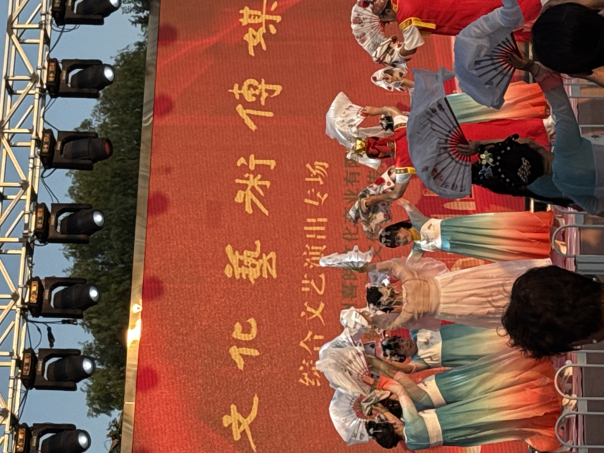 The image depicts an outdoor performance on a stage with a large red backdrop featuring golden Chinese characters. Several performers, predominantly women and girls, are on stage dressed in traditional Chinese attire. Many are holding decorative fans, some open and some closed, and appear to be engaged in a dance or theatrical presentation. The backdrop has text that reads "文化艺术传..", which translates to "Culture and Art Transmission...". Below this, smaller text indicates a "Comprehensive Cultural and Artistic Performance Special Session". The lighting rig above the stage is visible, with several black spotlights aimed downwards. The background shows trees and a clear sky, suggesting it is daytime. The scene appears to be part of a cultural event or festival. There is a partial view of an audience member in the foreground.