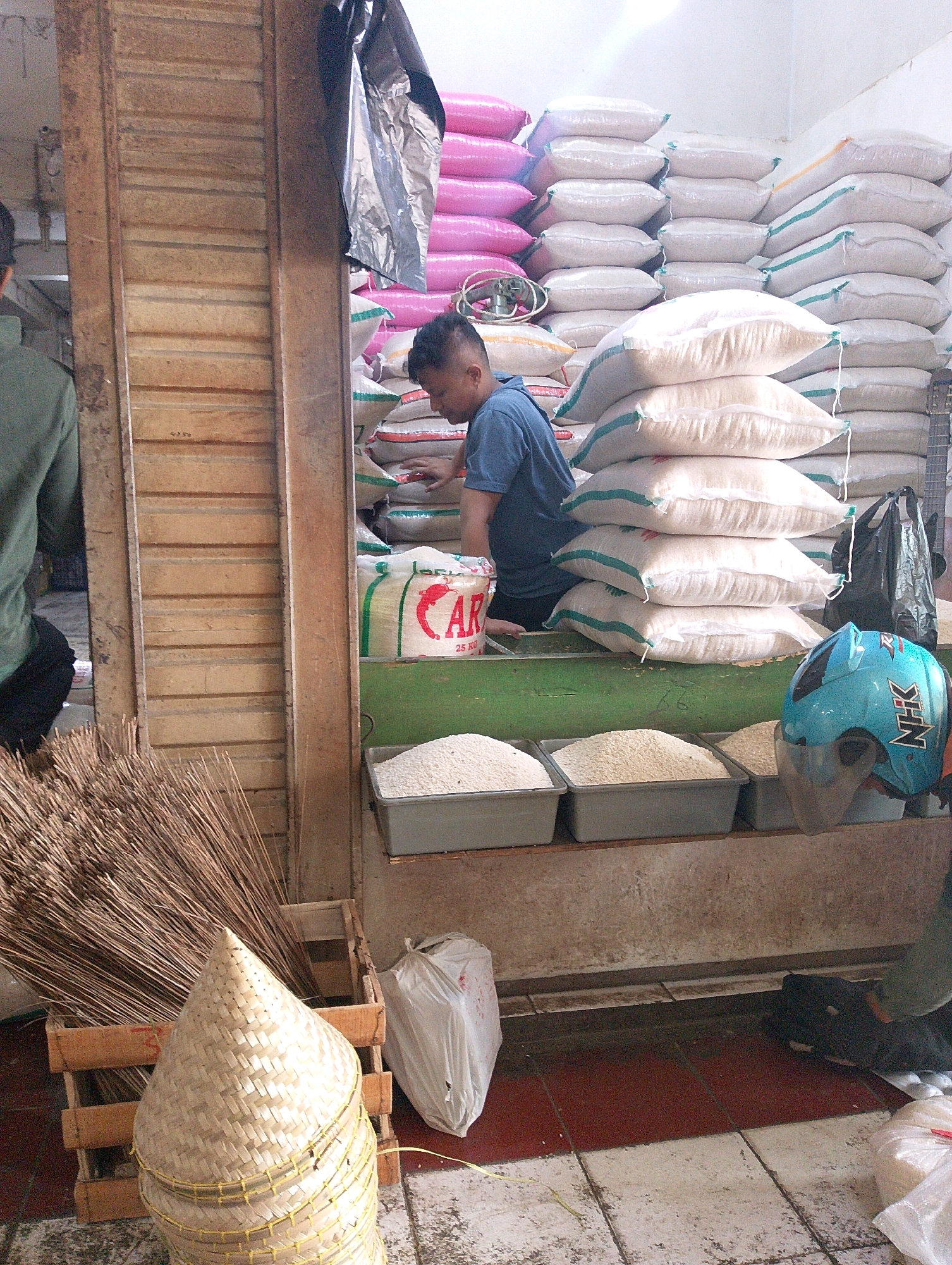 The image depicts a market stall in Lembang, Indonesia, likely a traditional market given the stacked sacks of rice and the presence of brooms. A man wearing a blue t-shirt is visible behind a counter, partially obscured by sacks of white rice. Several large sacks of rice, some with green and red stripes, are stacked high behind him. In the foreground, to the left, a large bundle of brown brooms is propped in a wooden crate, alongside a woven conical basket. To the right, a blue motorcycle helmet with white and black decals sits on a green surface. Three rectangular gray containers filled with white granular material, likely rice, are also present on the green surface. The floor is tiled with red and off-white squares. The overall scene suggests a commercial environment focused on the sale of rice and related goods.