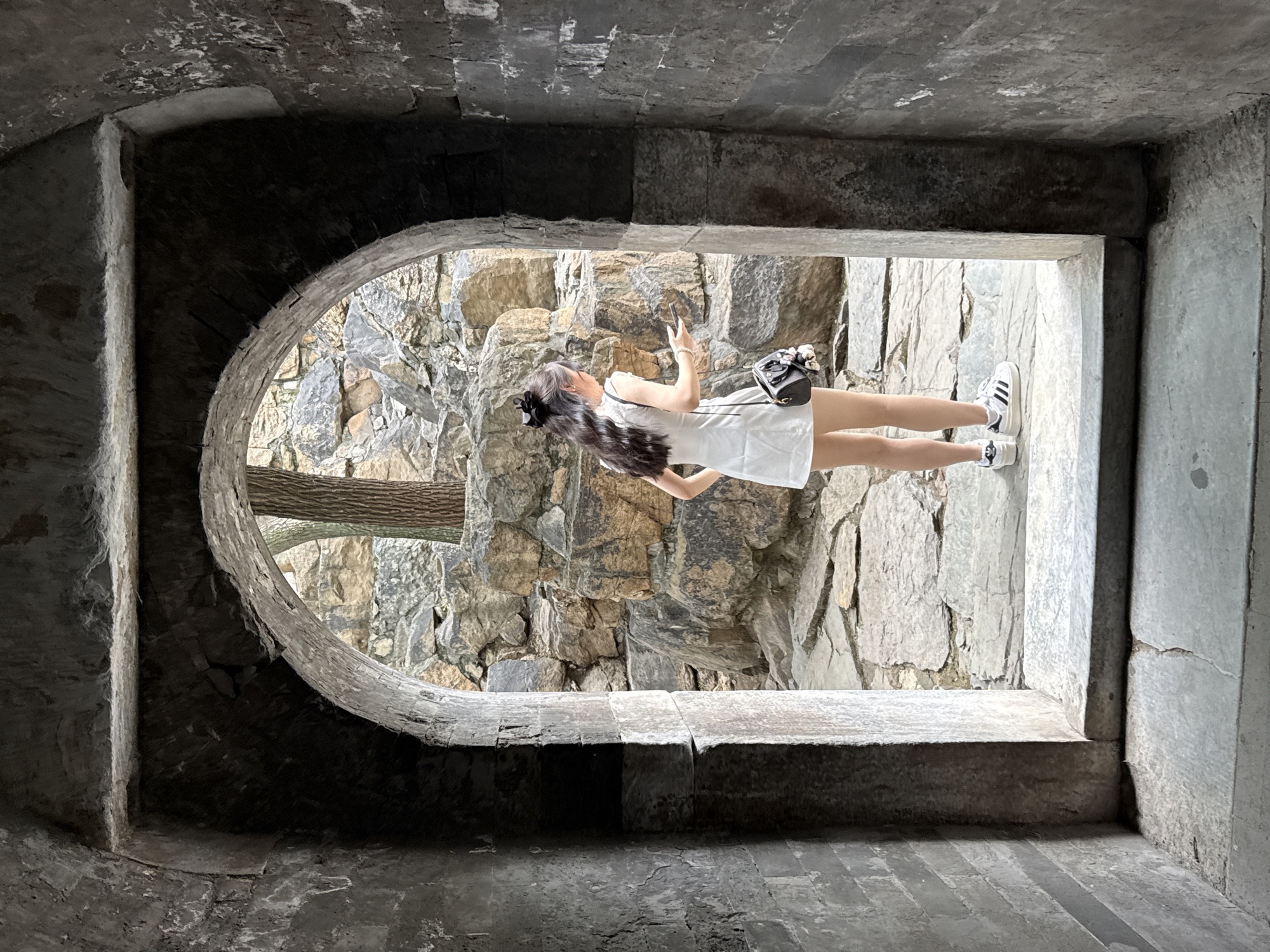 The scene depicts a female individual standing at the threshold of a stone archway, facing away from the viewer, in an outdoor setting within Haidian District, China.
The archway, constructed from dark grey, rough-textured stone or brick, frames the central subject. The interior surfaces of the arch and its supporting walls are deeply shadowed, revealing uneven textures and some lighter-colored patches, possibly efflorescence, particularly on the upper right side. The floor within the archway is composed of large, flat, dark grey stone slabs, with a distinct, raised stone threshold marking the transition to the exterior ground.
Beyond the archway, the environment is brightly lit and dominated by a rugged landscape of large, irregularly shaped natural rocks and boulders, ranging in color from grey to warm brown and ochre. A tree trunk with visible bark texture stands partially visible on the right side of the frame, behind the individual. The ground outside the archway is paved with irregular flagstones, creating a naturalistic pathway.
The individual is a female, seen from a three-quarter back view, positioned slightly to the right of the archway's center. She has long, dark, wavy hair, tied up at the crown with a black hair accessory, possibly a scrunchie or bow. She is wearing a sleeveless white mini-dress. A small, dark-colored handbag, adorned with white decorative elements (appearing as pompoms or flowers), is slung over her right shoulder. Her legs are bare, and she is wearing white athletic shoes with black stripes and laces. Her right hand is holding a white-cased smartphone, with her fingers positioned on the screen, indicating interaction with the device. Her gaze is directed towards the right, into the rocky background, presumably at the phone screen.