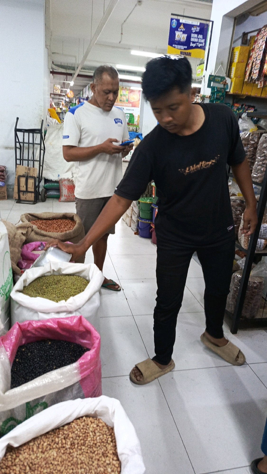 In a brightly lit indoor marketplace in Ponorogo, Indonesia, two men are engaged in a transaction near sacks of bulk grains. The man on the left, wearing a white t-shirt with a blue collar and grey shorts, holds a smartphone. The man on the right, dressed in a black t-shirt and black pants, is reaching into a large white sack filled with green beans with a metal scoop, preparing to weigh or package them. Visible in the foreground are three large sacks of different legumes: one with black beans, another with what appears to be mung beans, and a third at the very bottom with smaller, reddish-brown beans. In the background, shelves are stocked with various packaged goods, and a shopping cart is visible. The floor is tiled.