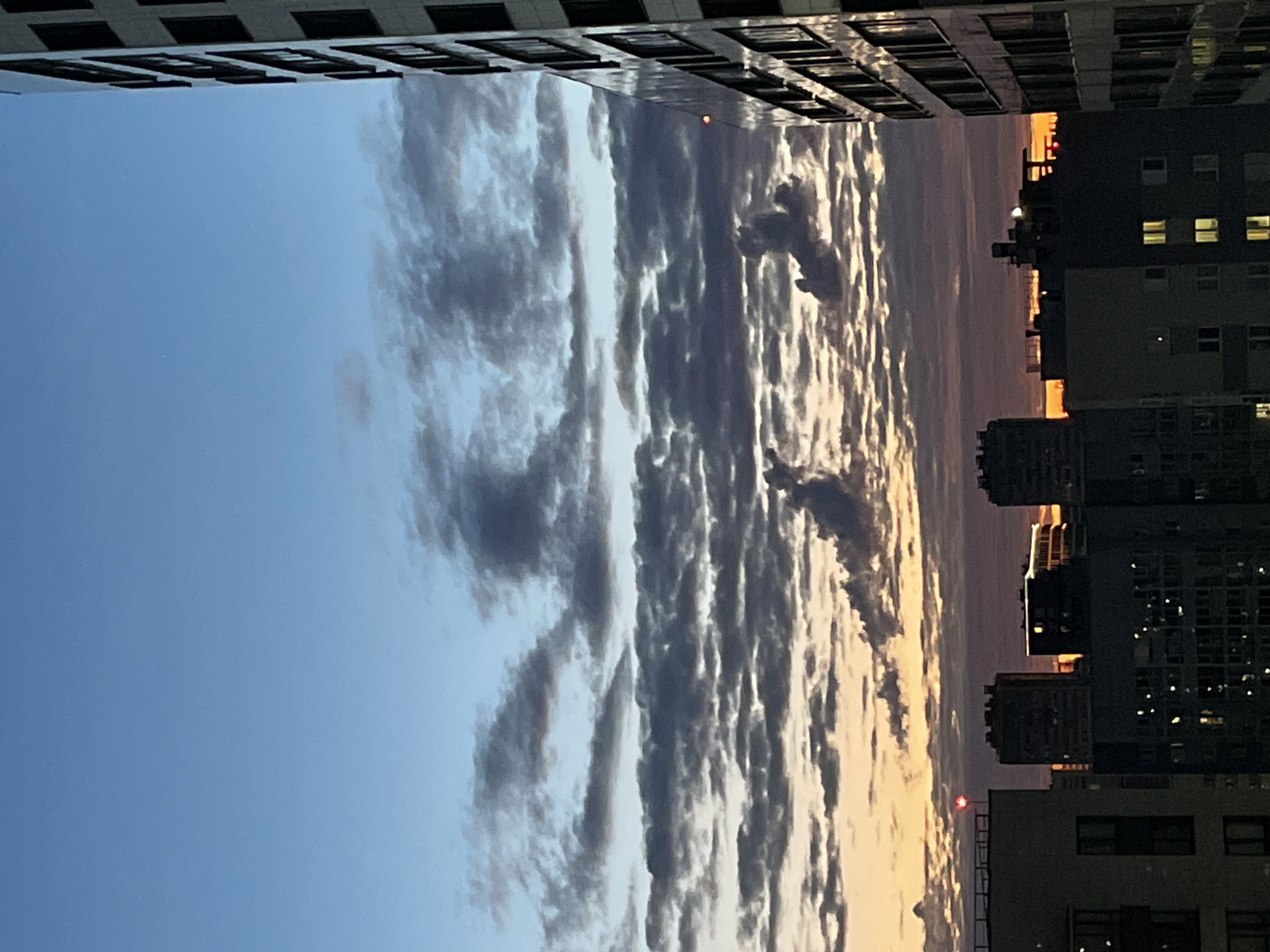 This is a photograph of a city skyline at dusk, likely taken from a high vantage point. The sky is a gradient of blue and purple, with scattered clouds that are illuminated by the setting sun. The clouds are dark and moody, with some streaks of orange and pink visible at the horizon.
In the foreground, several modern apartment buildings are visible. These buildings are tall and rectangular, with many windows. The building on the right is particularly prominent, its facade reflecting the colors of the sky. The buildings in the background are silhouetted against the sky, and some have lights on in the windows. There are no people visible in the image. The overall impression is one of an urban landscape at the end of the day, with a dramatic and somewhat somber sky. The photo was taken in the city of Unknown, Russia.